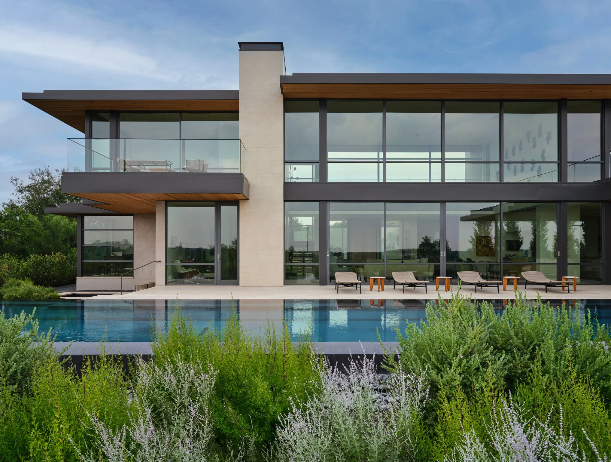 Contemporary limestone residence with expansive glass walls overlooking an infinity-edge pool. Deep roof overhangs, wood soffits, and restrained landscaping frame views toward the surrounding Texas Hill Country.