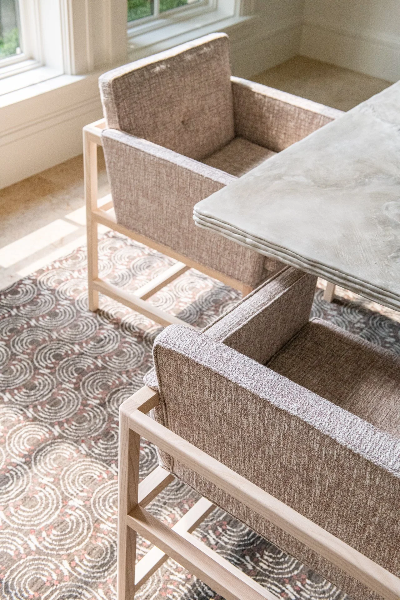 Upholstered dining chairs with pale wood frames by Bassam Fellows surrounding a sculptural stone dining table by BAILEY FONTAINE STUDIO in a warm contemporary dining room dappled in sunlight.
