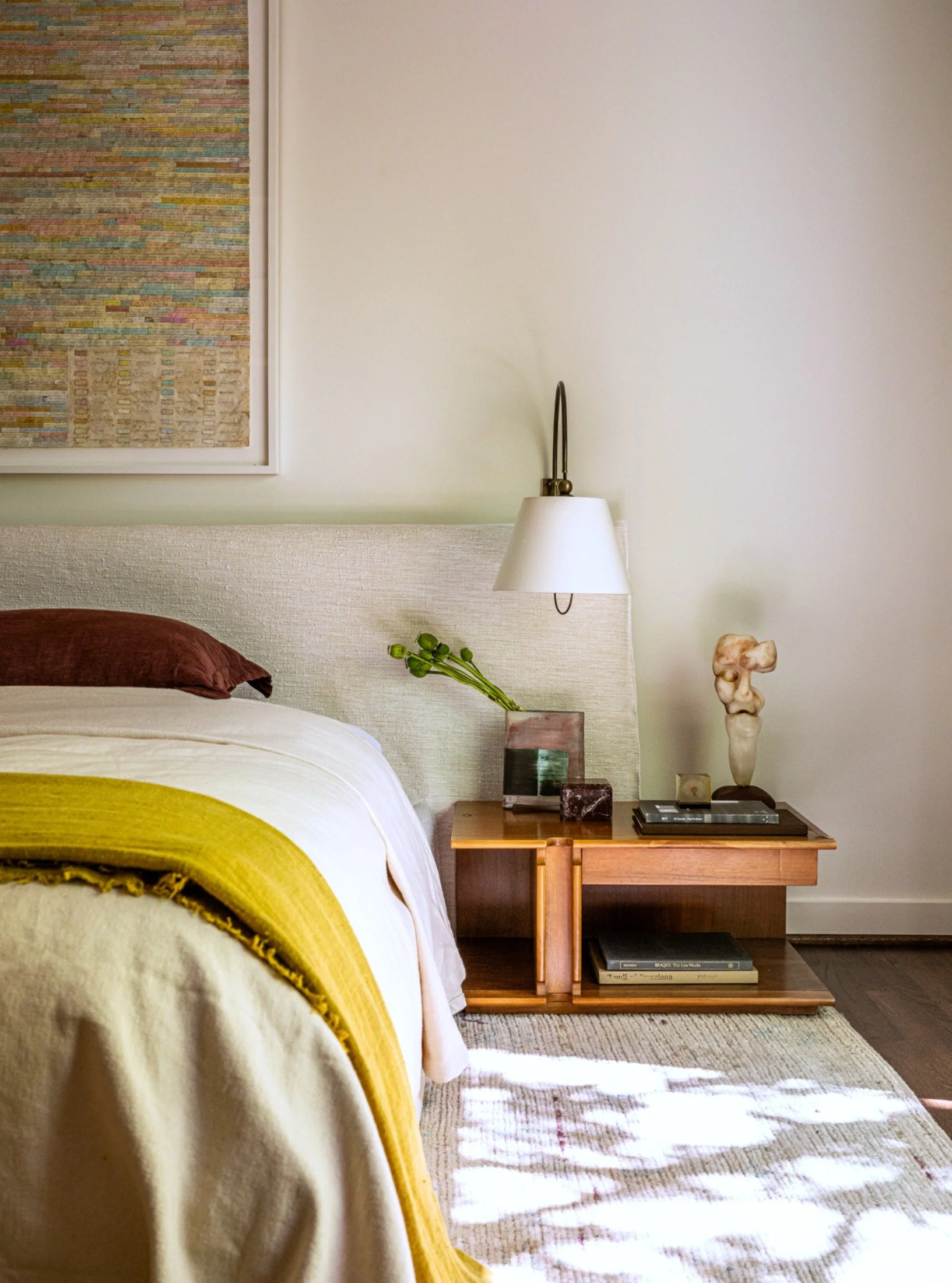 Contemporary bedroom with upholstered headboard, vintage wood nightstand by Bernini, and layered textiles, styled with sculptural objects and warm natural materials.
