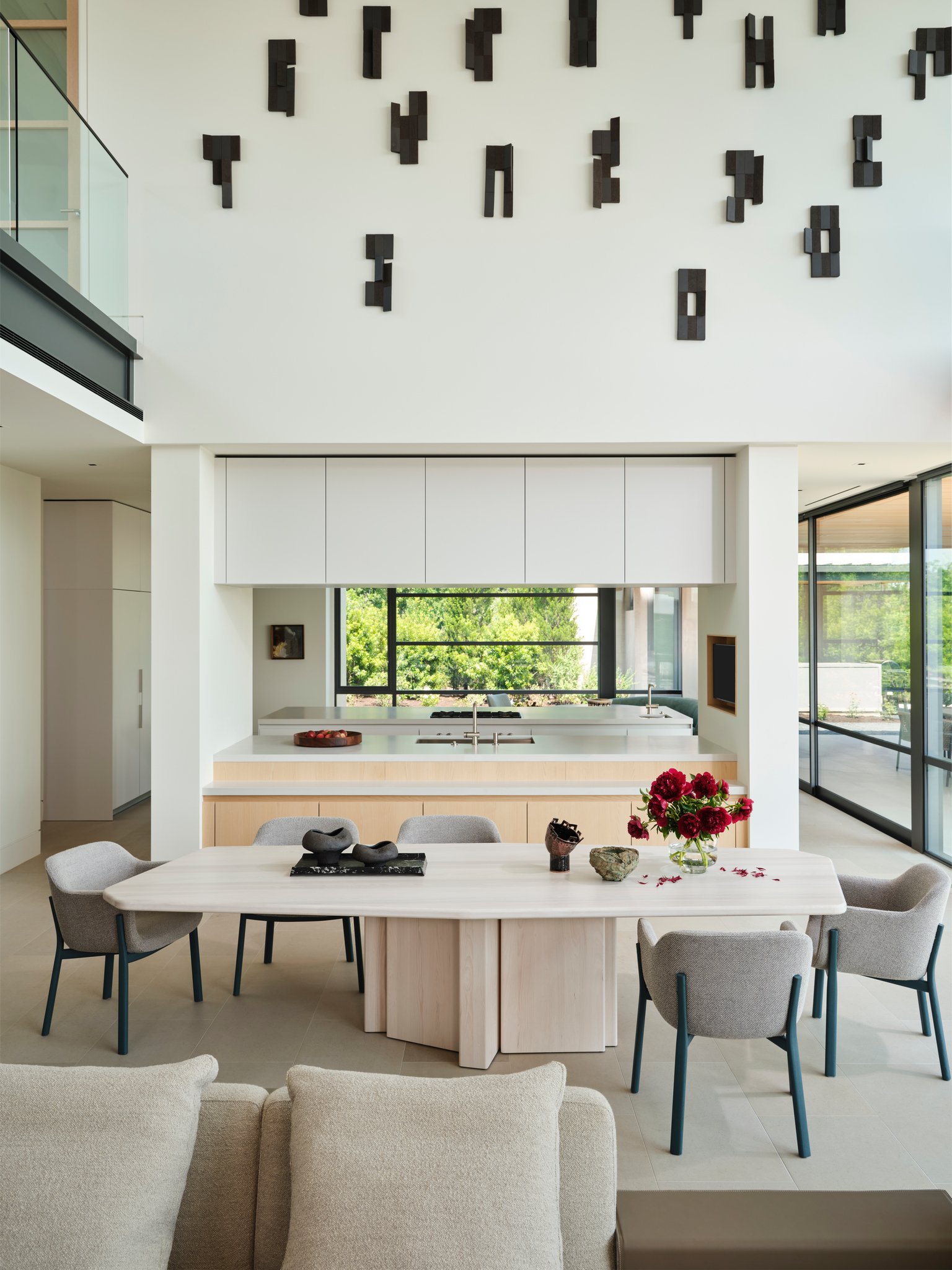 Minimal dining space with pale wood table and contemporary chairs positioned between kitchen and living areas. A dramatic double-height wall features a large abstract wall sculpture installation.  simon johns table, sculptures by denis castaing