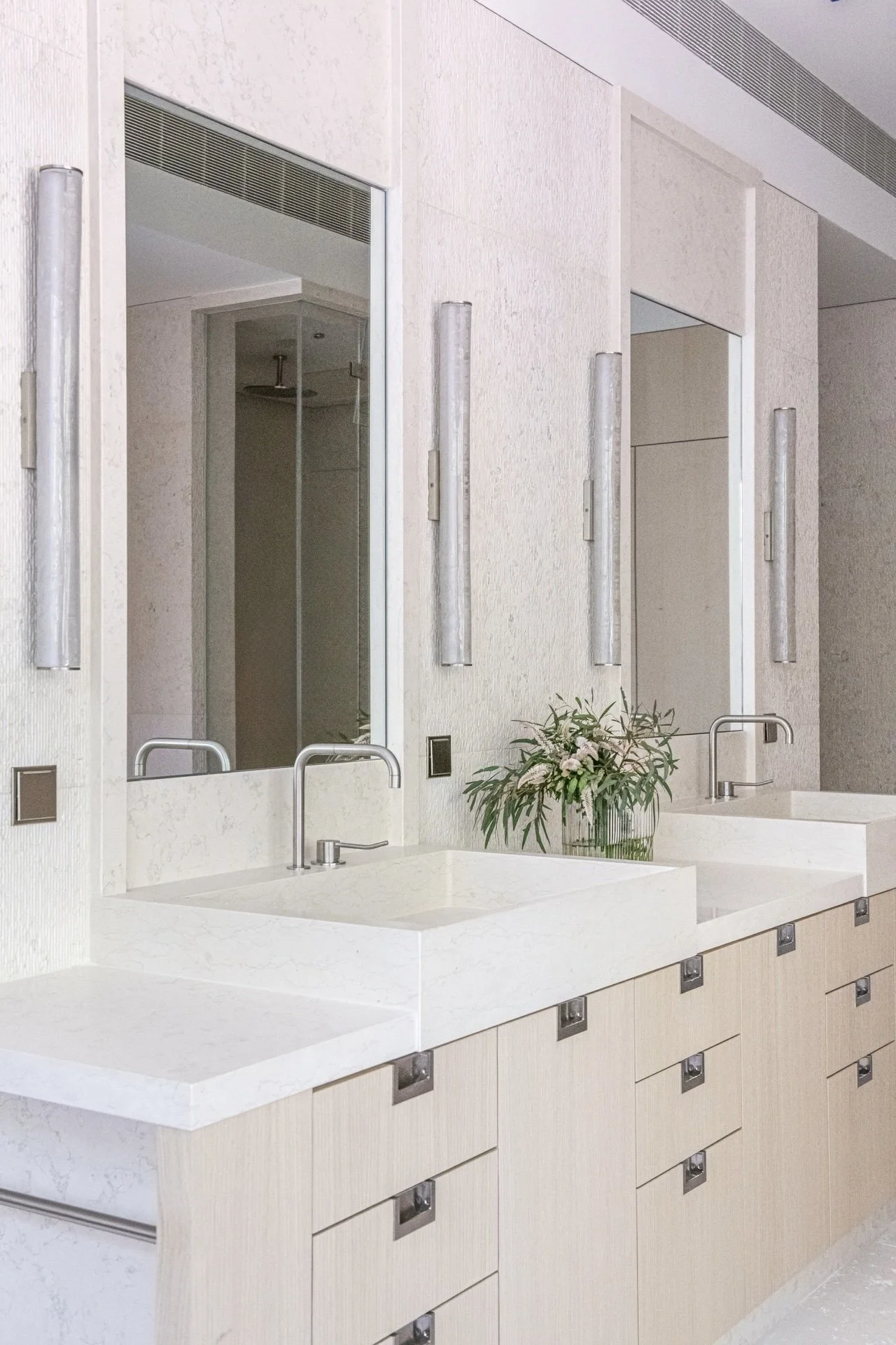 Double vanity bathroom with integrated stone sinks, pale wood cabinetry, textured Bianco Perlino walls, and understated modern elegance.