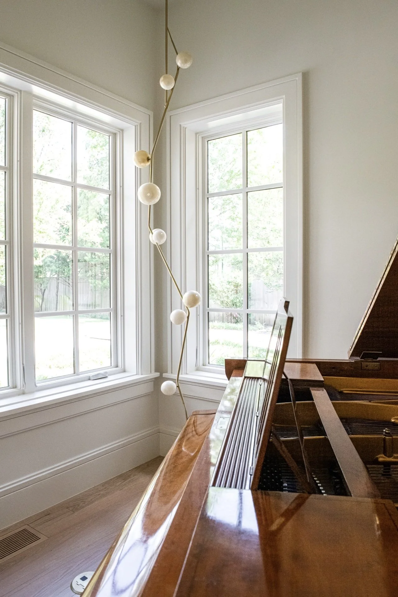 Grand piano corner with sculptural brass pendant light by Lindsay Adelman, tall windows, and soft natural light in a refined residential music room.