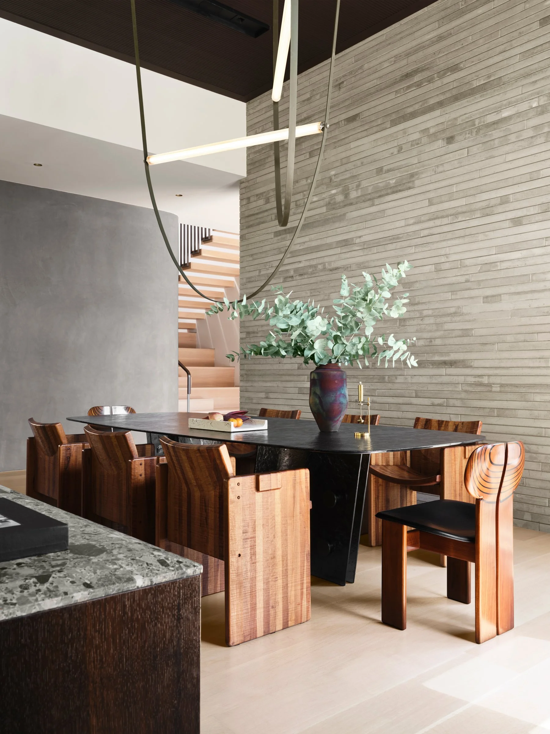 Dining space with solid wood chairs, sculptural table, and contemporary lighting, set against textured masonry walls in a modern home. Vintage Giuseppe Rivadossi Hand Carved Unique Dining Chairs. Vintage Tobia and Afra Scarpa ‘Artona’ chairs at head 