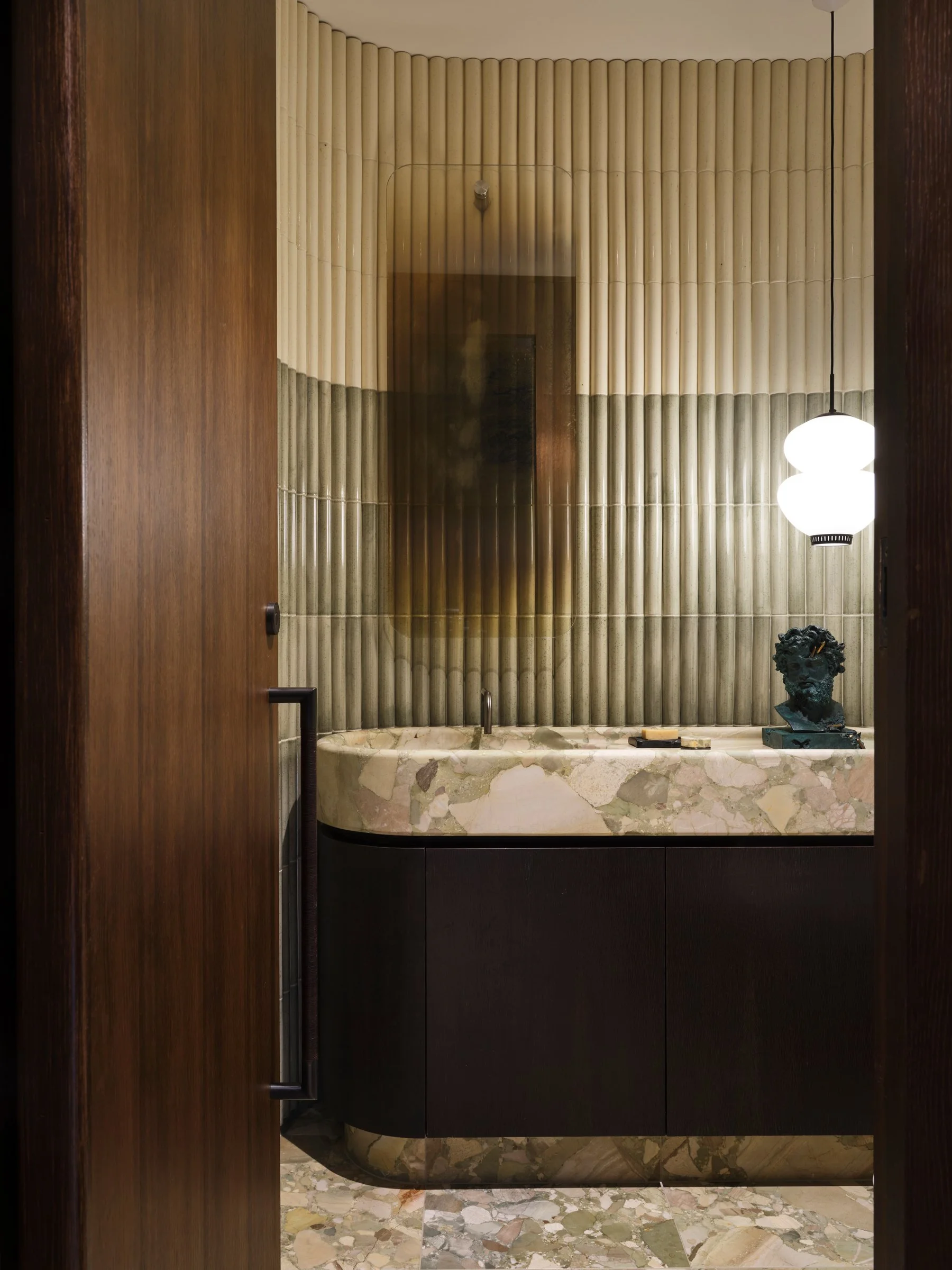 Powder room with fluted ceramic wall tile and custom green ceppo vanity, layering tonal variation and sculptural surface articulation.