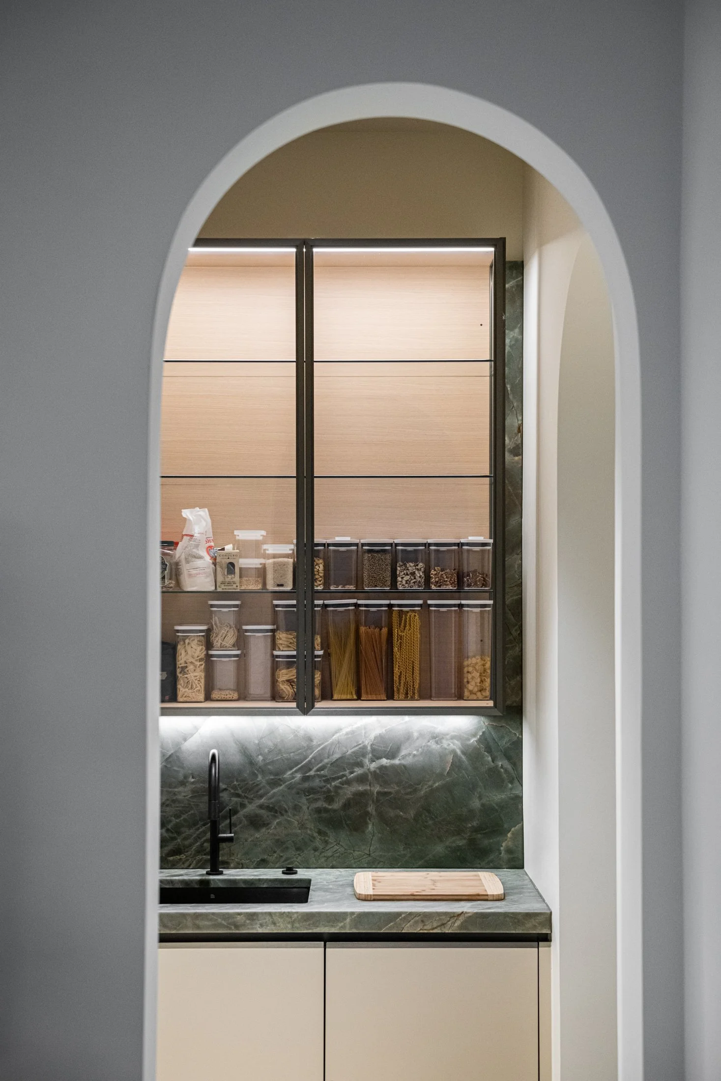 A softly framed pantry reveals stone counters, illuminated shelving, and organized everyday storage. Functional utility is elevated through architectural proportion and carefully resolved material continuity.
