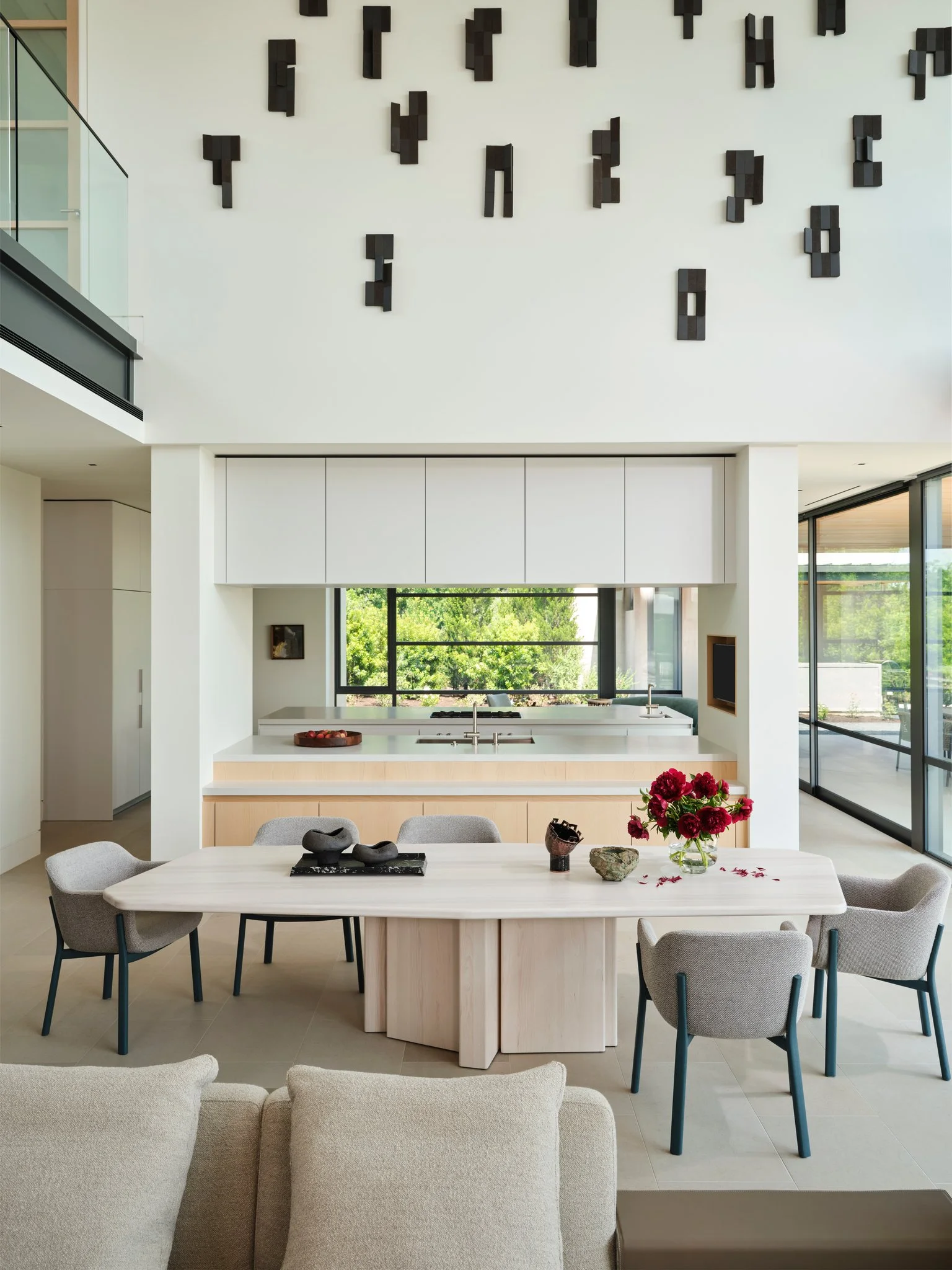 Minimal dining space with pale wood table and contemporary chairs positioned between kitchen and living areas. A dramatic double-height wall features a large abstract wall sculpture installation.  simon johns table, sculptures by denis castaing