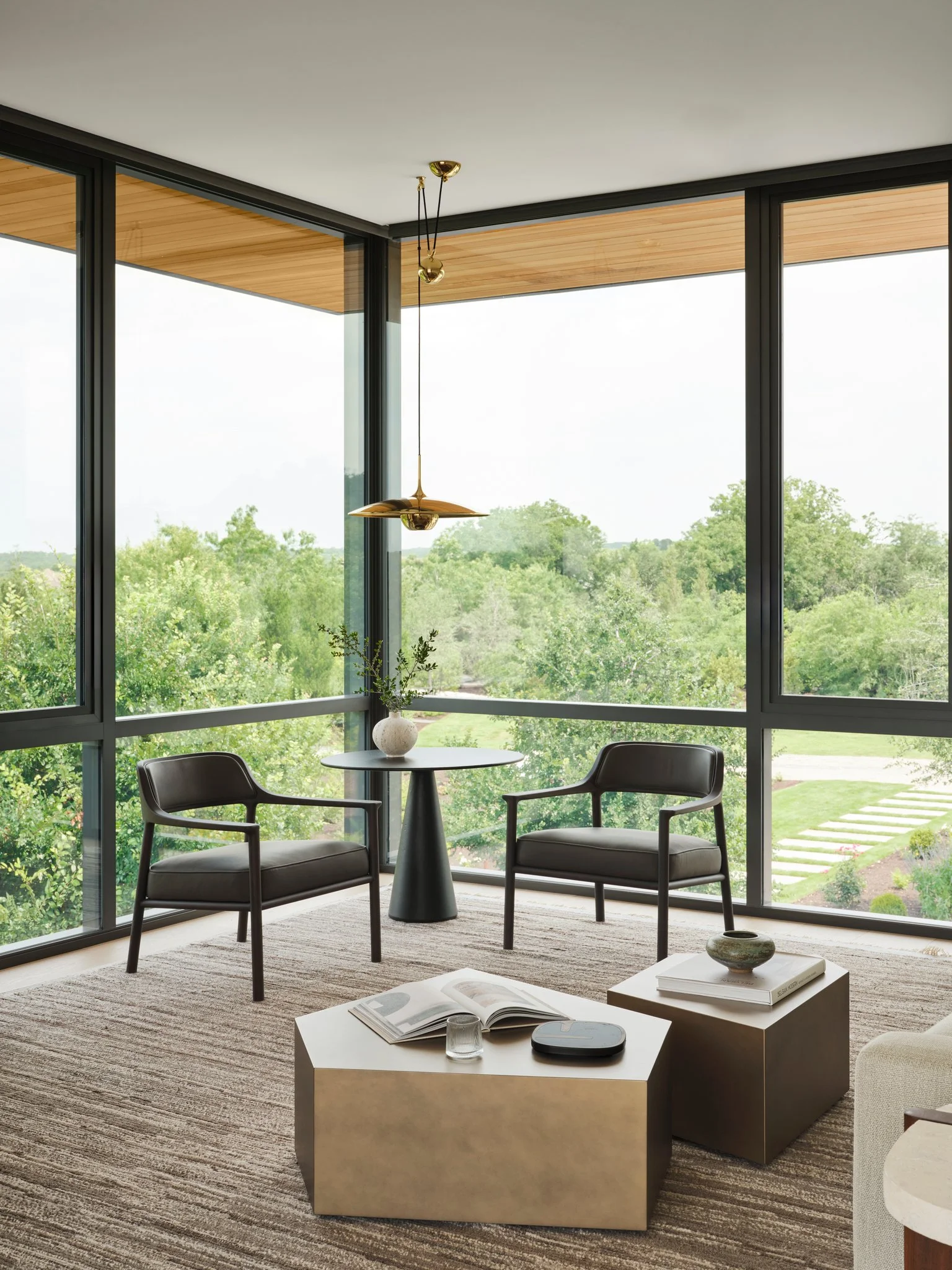 Quiet sitting area framed by floor-to-ceiling glass overlooking Texas Hill Country landscape. Sculptural metal coffee tables, leather Molteni lounge chairs, and warm wood soffit create a refined modern interior. lamp by florian schulz