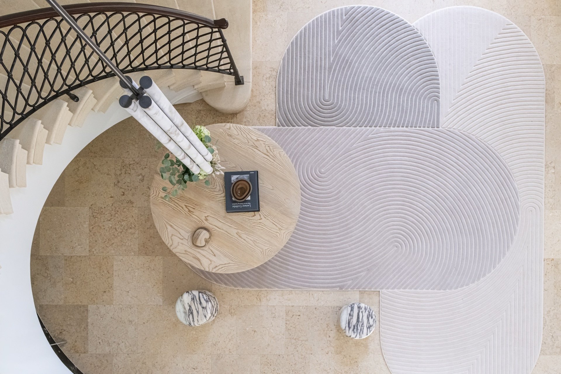Overhead view of a grand foyer featuring curved staircase, custom rug by Joshua Rice   Design, marble stools by Michael Verheyden, and sculptural oak center table by Casey McCafferty beneath alabaster and bronze architectural lighting.