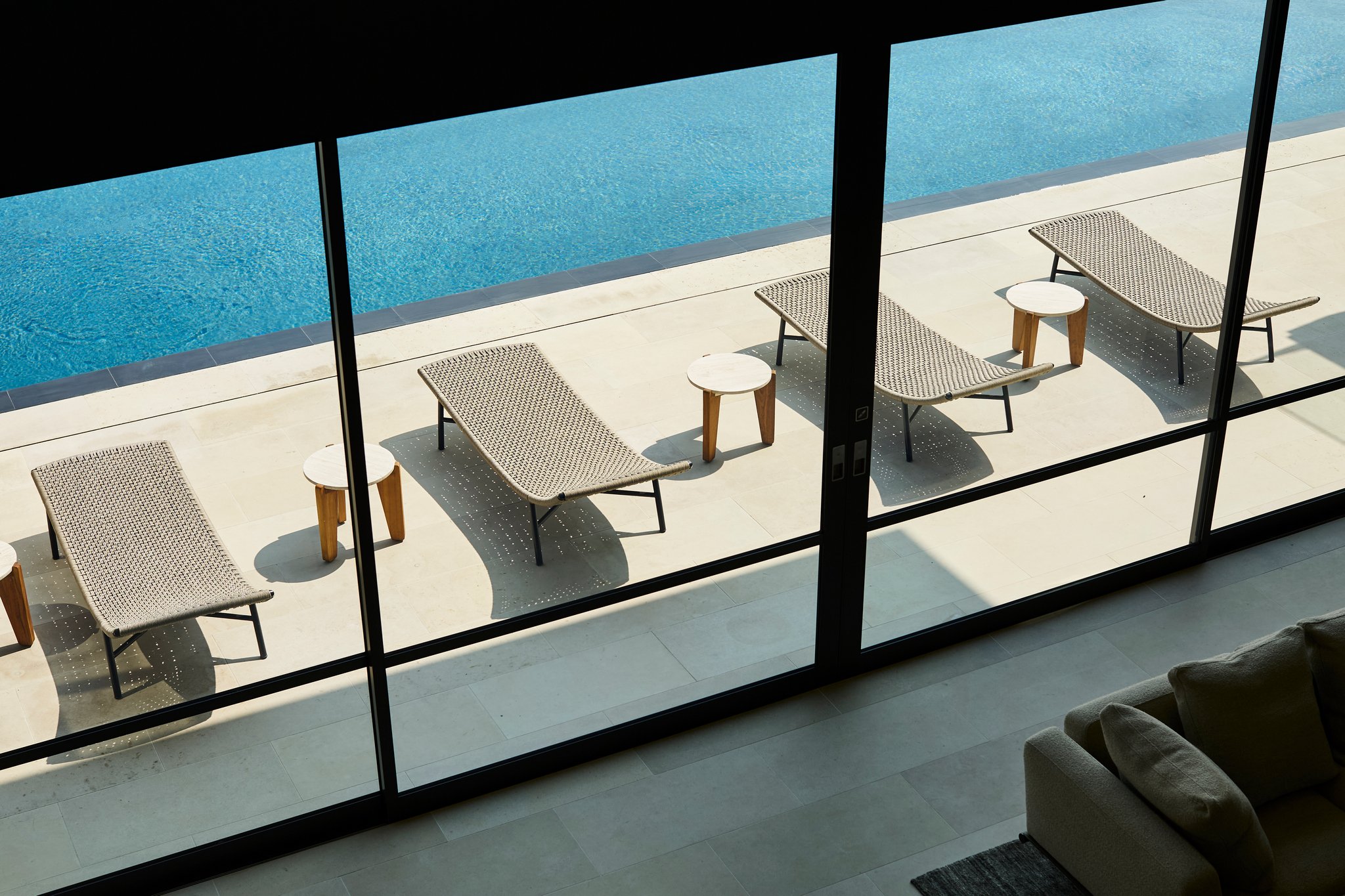 View from interior toward the pool terrace with woven lounge chairs and small wood tables arranged beside a long swimming pool and expansive landscape views. furniture by jader Almeida
