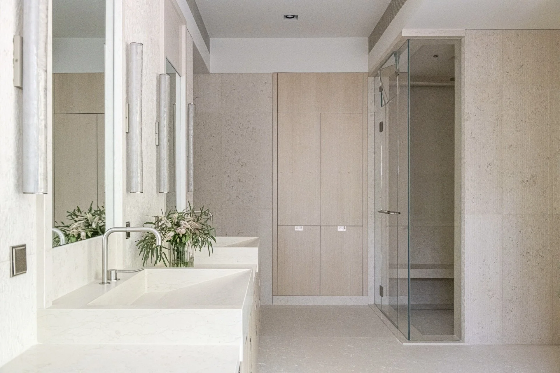 Spacious primary bathroom with double stone vanities, glass shower enclosure, pale oak cabinetry, and warm minimalist interior architecture.