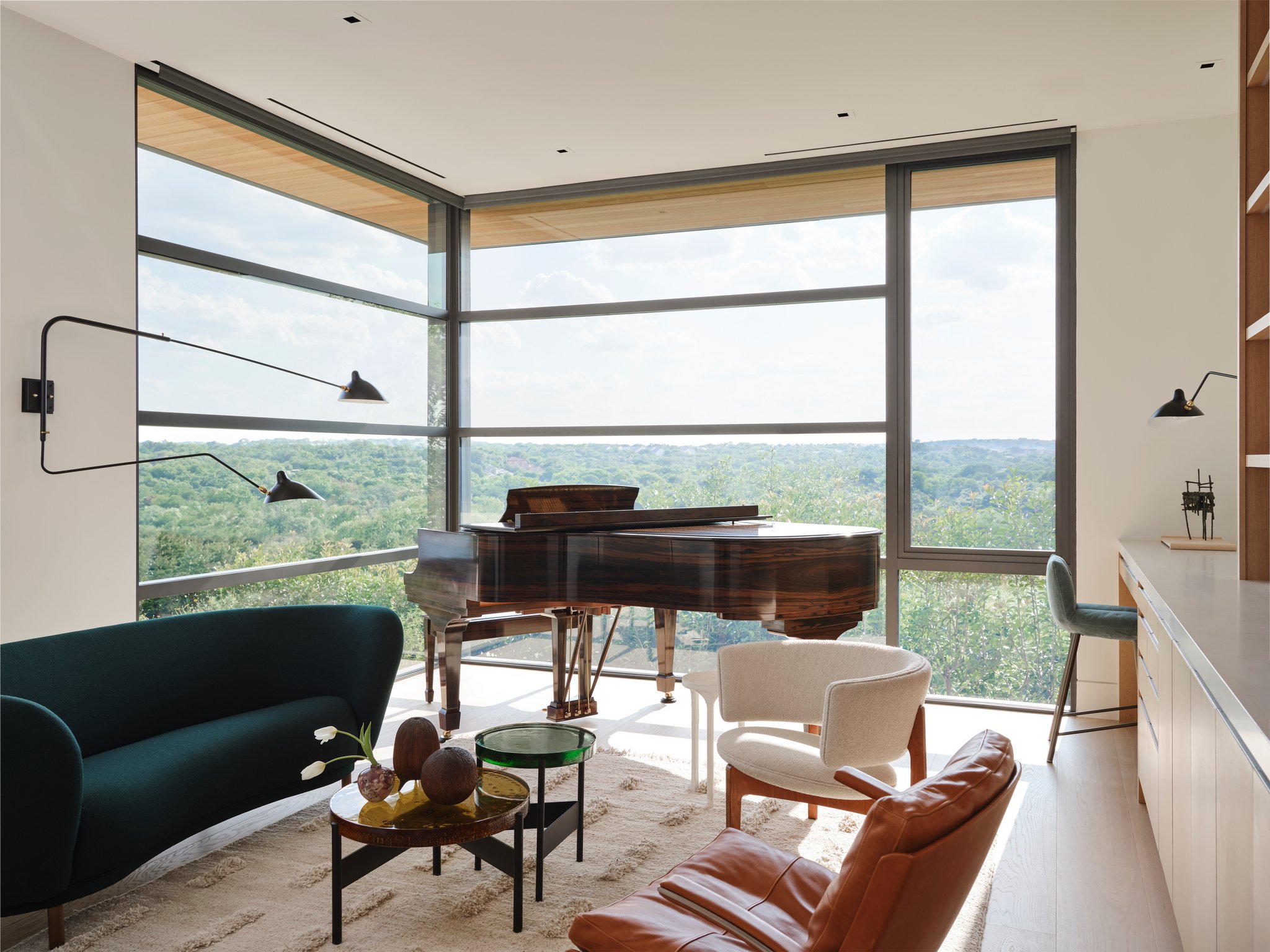 Sunlit library room anchored by a grand piano and surrounded by floor-to-ceiling windows overlooking wooded landscape. Curated seating, sculptural tables, and gallery lighting create a calm architectural interior. massproductions, fabricius kastholm 