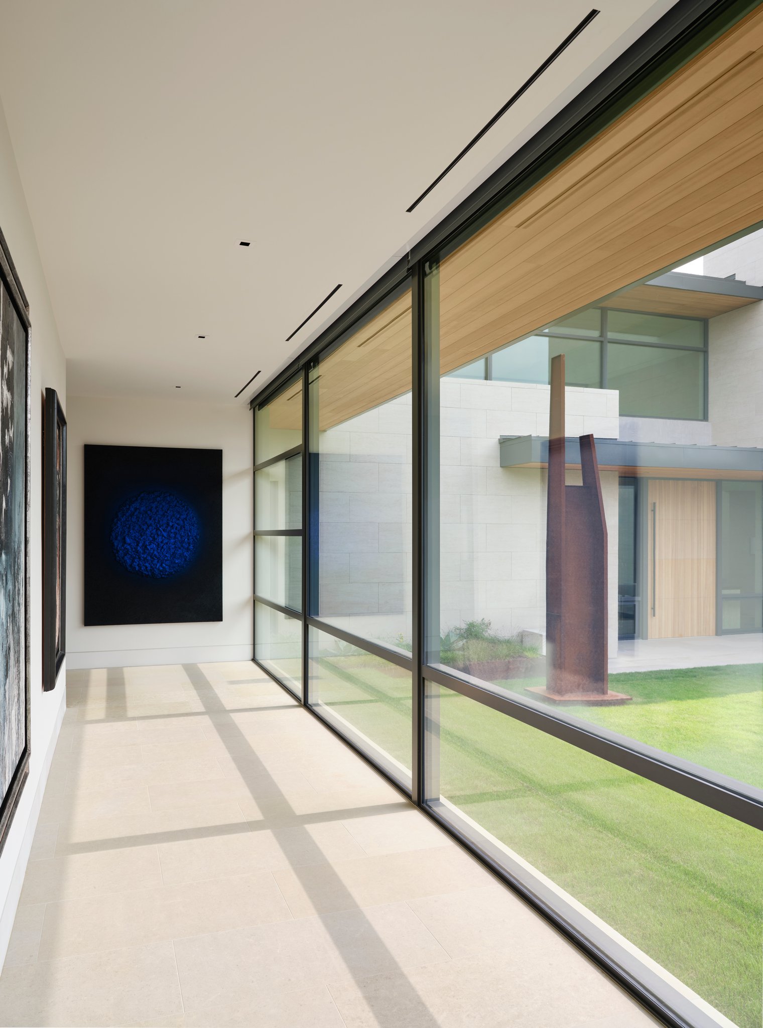 Long gallery hallway with floor-to-ceiling glazing overlooking courtyard sculpture and garden. Natural light washes across stone flooring and contemporary artwork.  sculpture by jonathan Nesci Samuel DeJong Antoni Clave