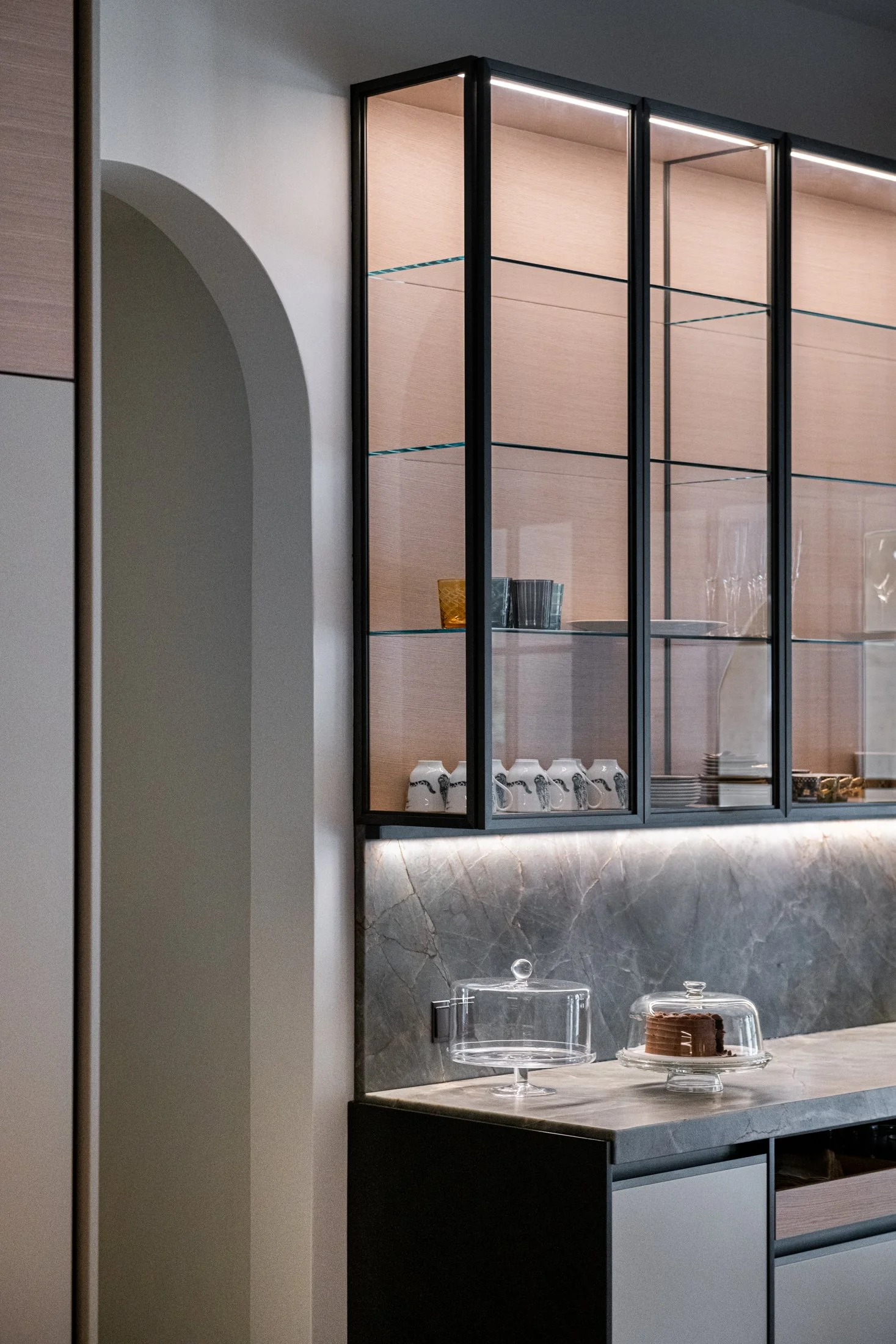 Built-in bar cabinet with illuminated glass shelving, stone backsplash, and tailored millwork in a sophisticated modern luxury kitchen by Vincent Van Duysen for Dada.