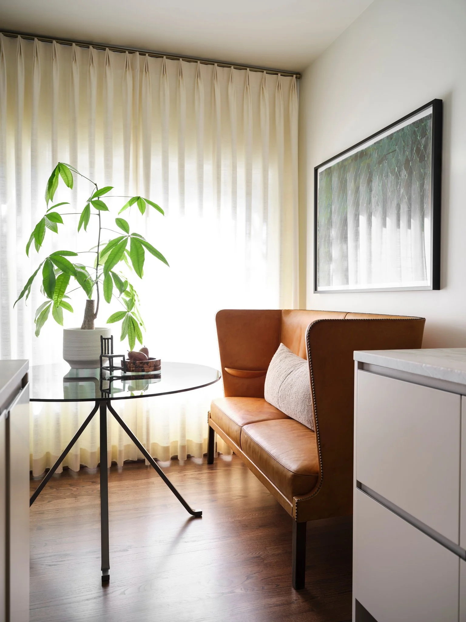 Modern breakfast nook with rare vintage leather sofa by Frits Henningsen, round glass table by Enzo Mari, and sheer drapery, featuring warm wood flooring and soft natural light in a contemporary residential interior.