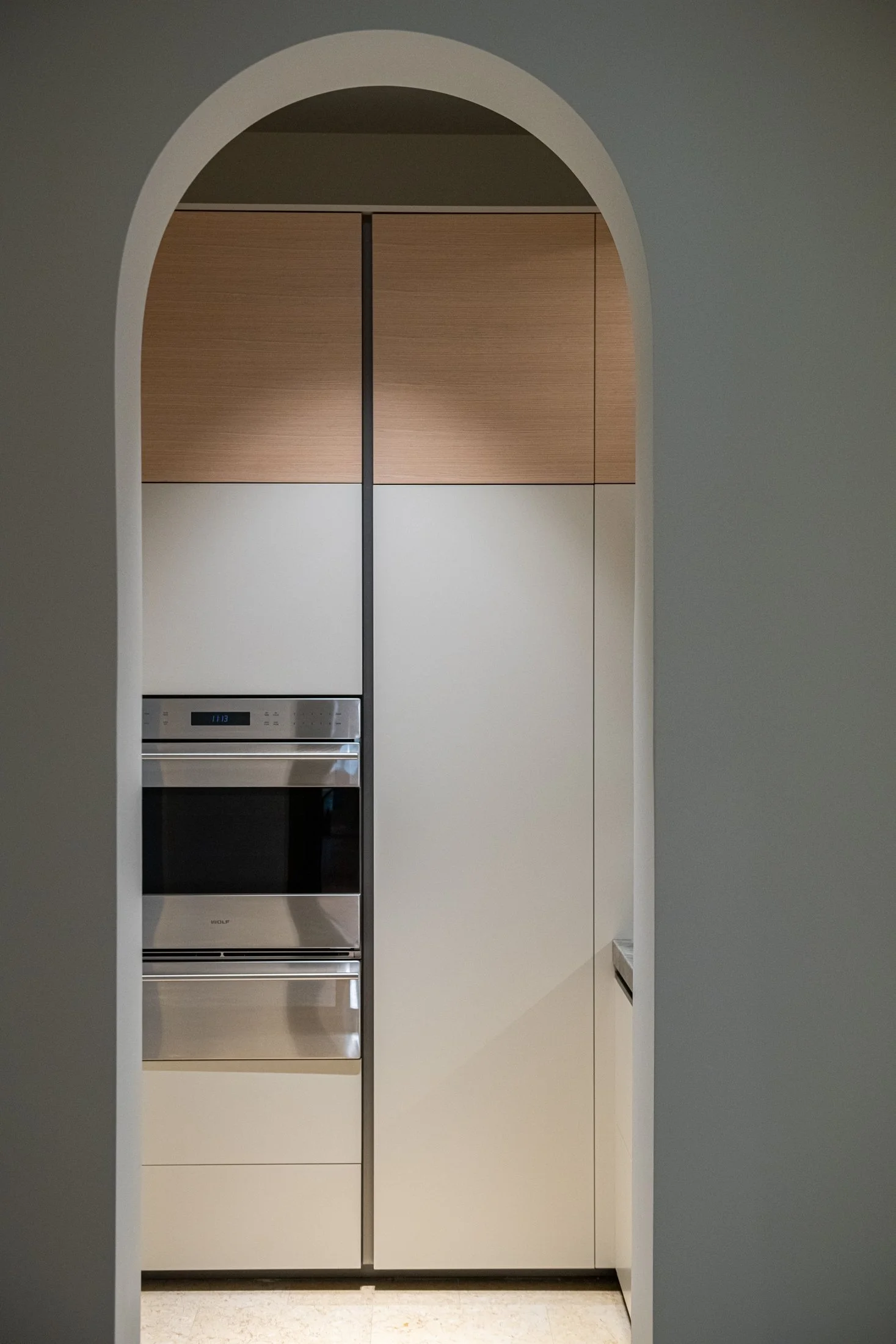 A softly framed arched opening introduces custom millwork and integrated appliances, balancing classical architecture with contemporary restraint. Warm oak and pale lacquer create a calm, tailored composition.