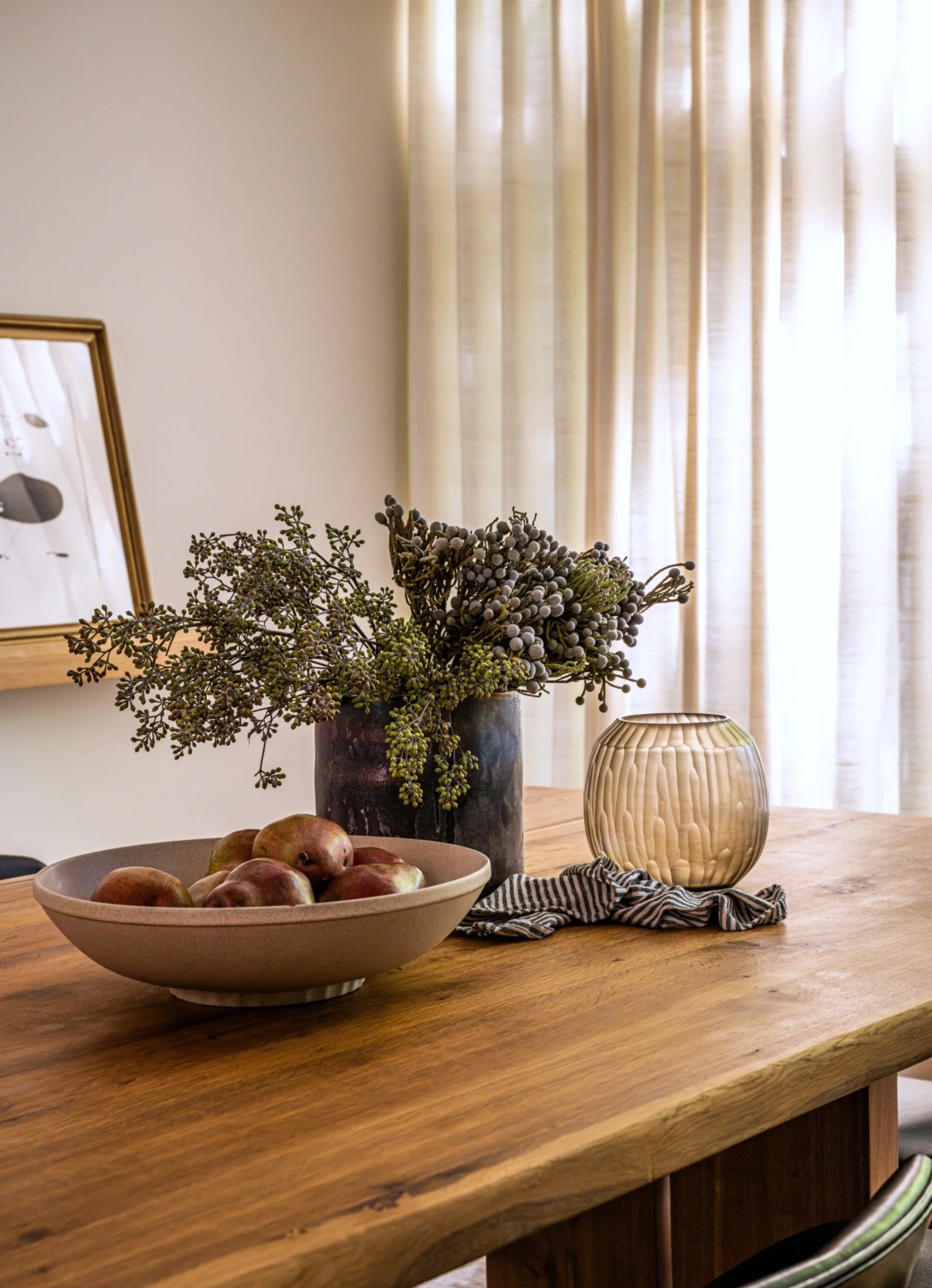 Solid wood dining table styled with ceramic bowl, fruit, and sculptural vessels, set against linen drapery in a material-driven modern residential interior.