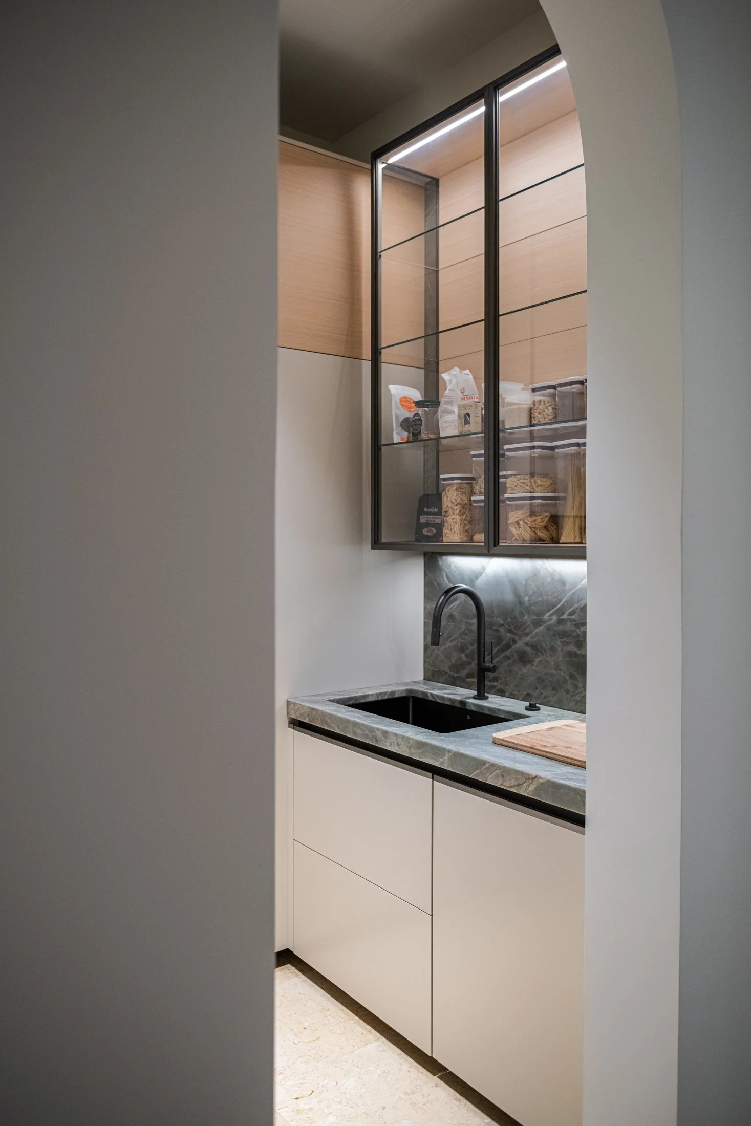 A compact service pantry with stone counters, blackened steel framed glass cabinetry, and integrated lighting. Functional storage is elevated through precise detailing and a restrained material palette.