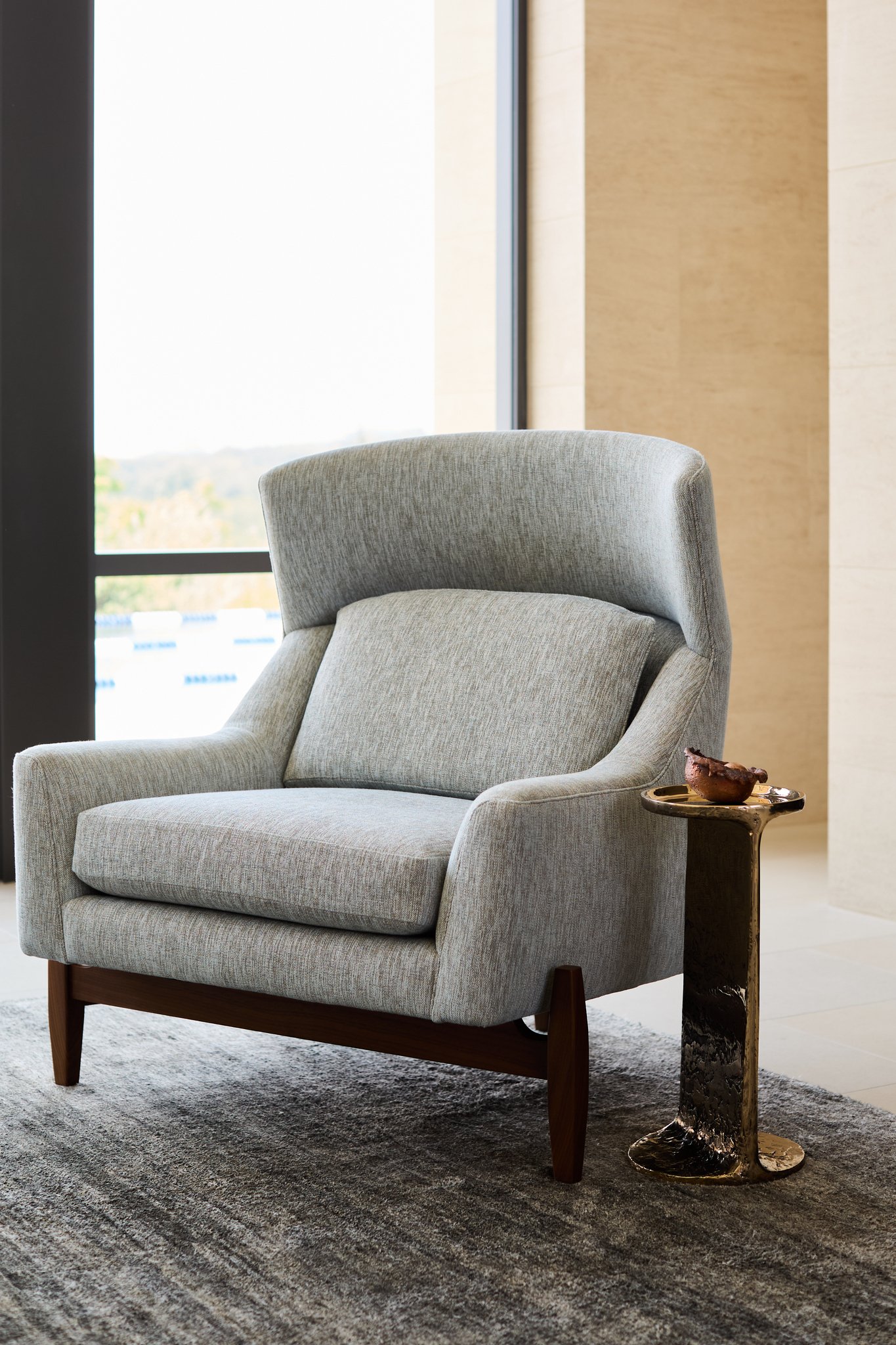 Detail of a soft upholstered lounge chair paired with a sculptural bronze side table beside a large window overlooking the landscape. Jens risom chair and refractory studio table
