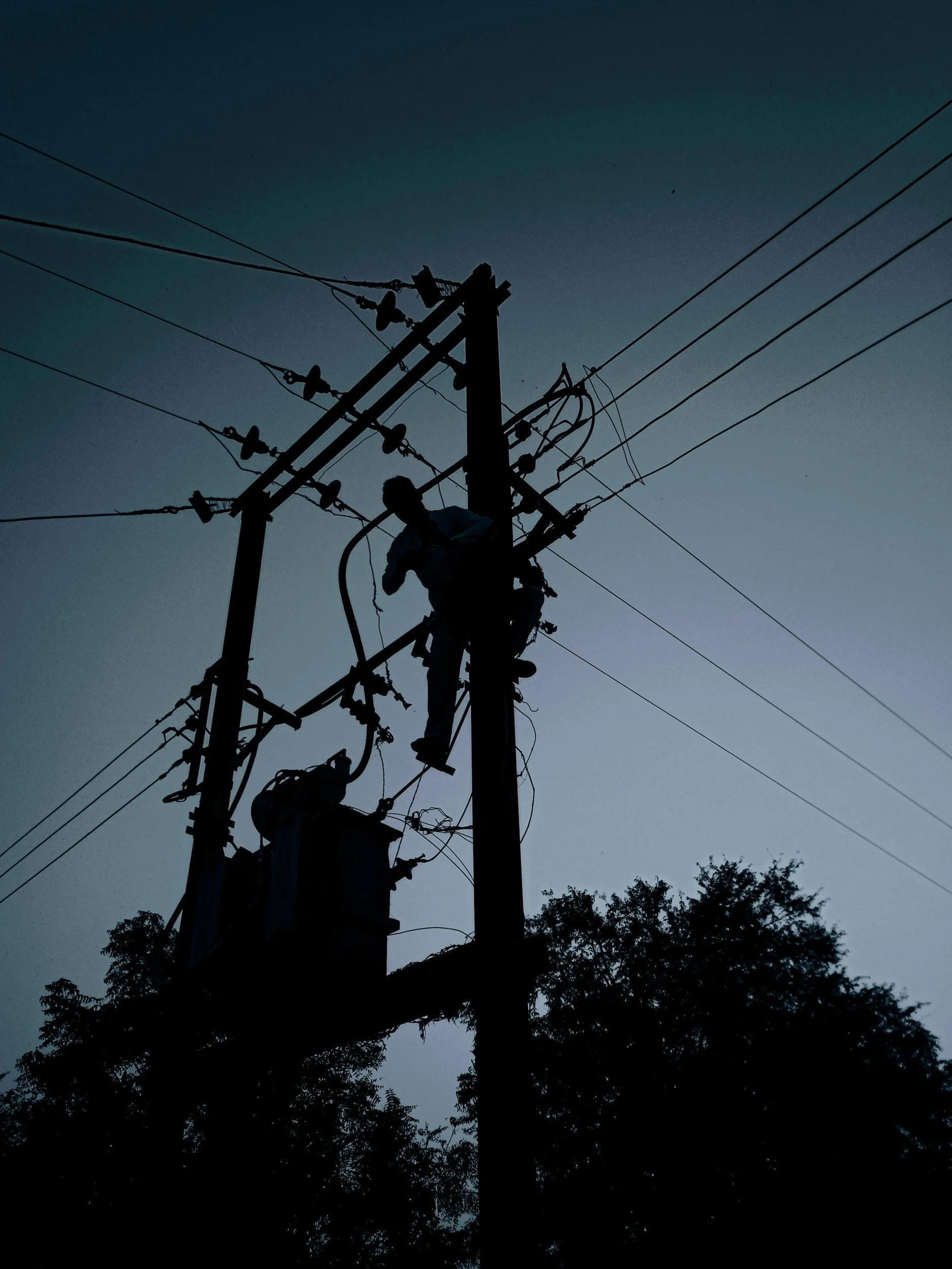 Silhouette of a utility worker repairing power lines on a telephone pole during dusk with trees in the background.