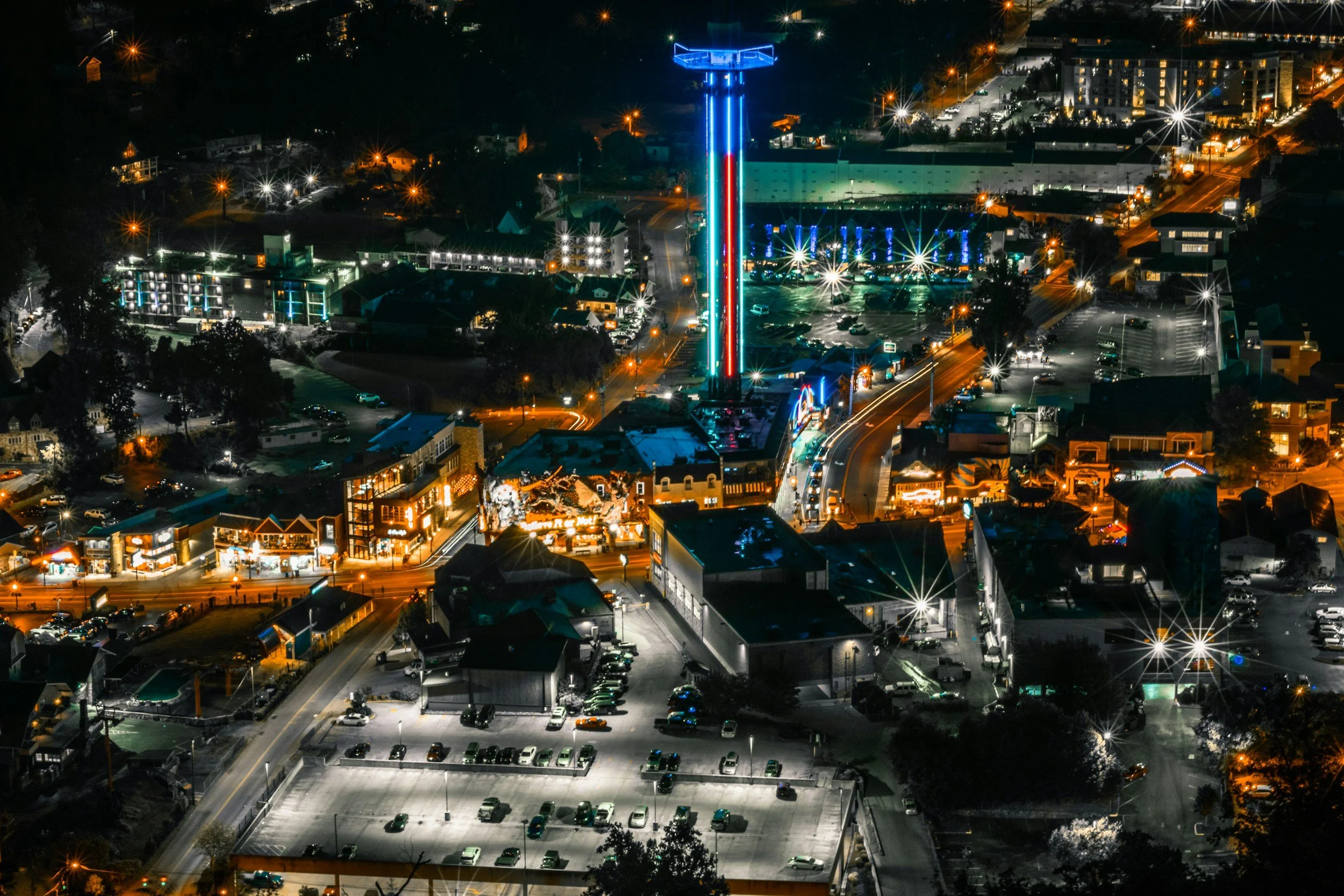 Night aerial view of a city street with illuminated buildings, neon signs, and a tall ride or tower with colorful lights.