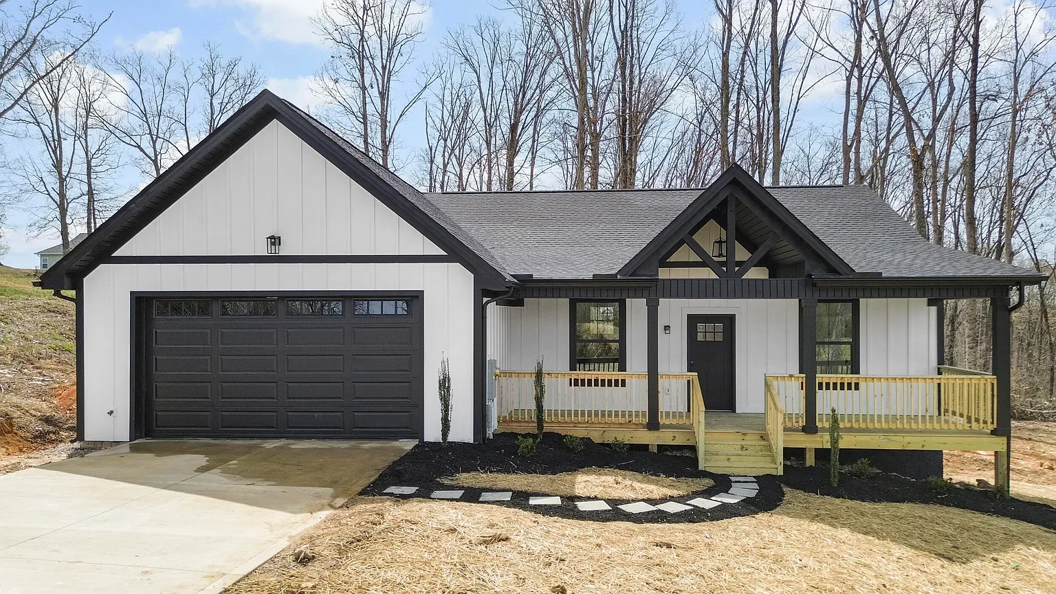 Newly built modern house with a white exterior, black accents, a gray roof, a front porch with a small staircase, and a attached garage, surrounded by a yard with mulch and stone walkway, with leafless trees in the background.
