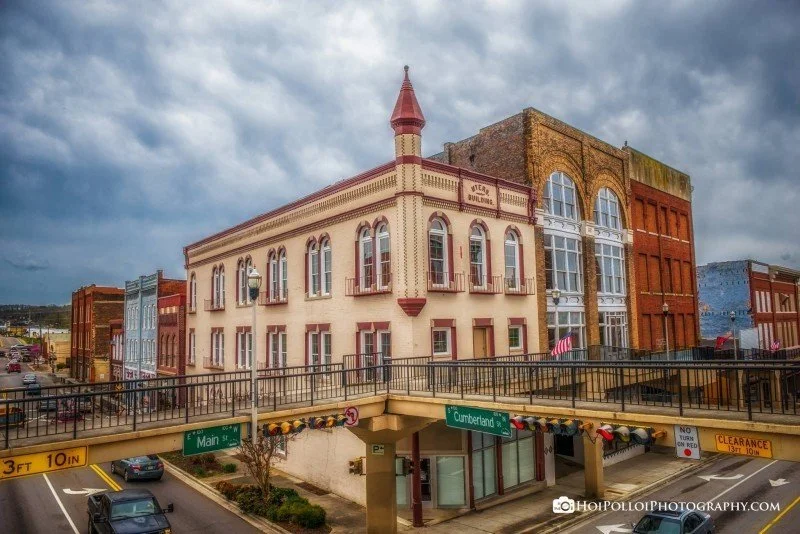 A historic downtown street scene with colorful, old-fashioned buildings, a bridge with street signs for Main Street and Cumberland Street, and cloudy skies overhead.