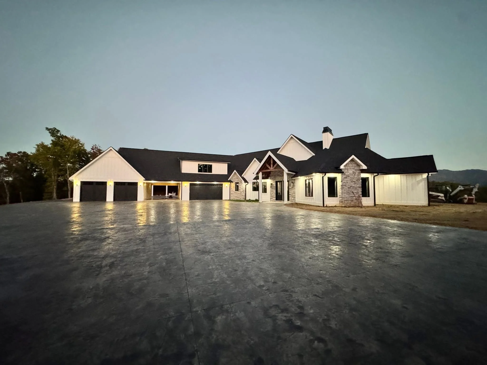 Modern house with a large driveway, exterior lights, and a dark roof, during dusk or early evening, with a cloudy sky and trees in the background.