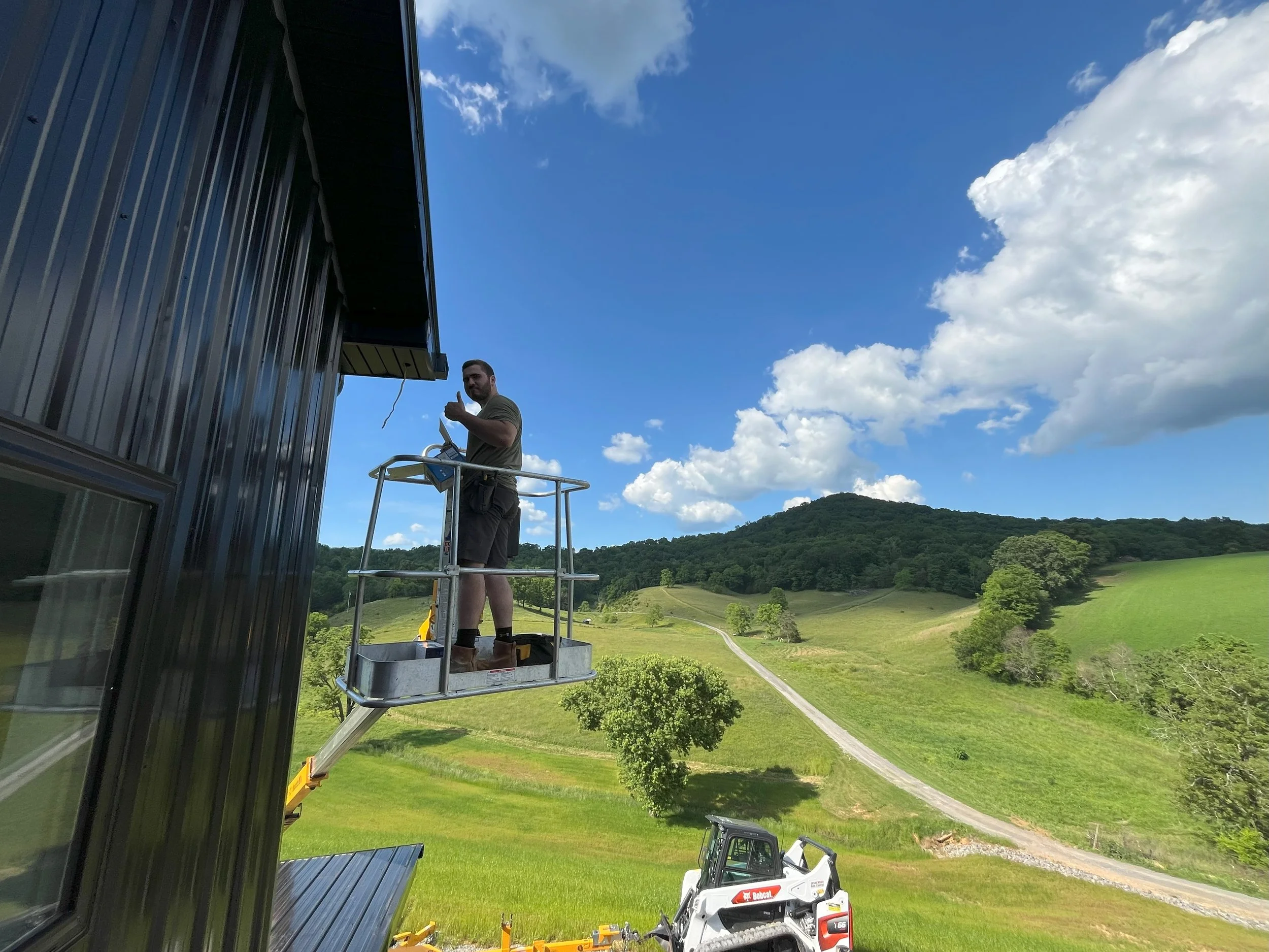 Man standing in a cherry picker work platform fixing or inspecting the side of a black metal building against scenic green fields and hills under a partly cloudy sky.