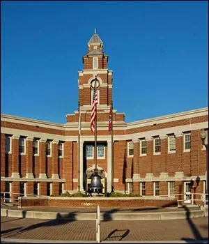 A historic brick building with a clock tower and a flagpole flying an American flag, with a clear blue sky in the background.