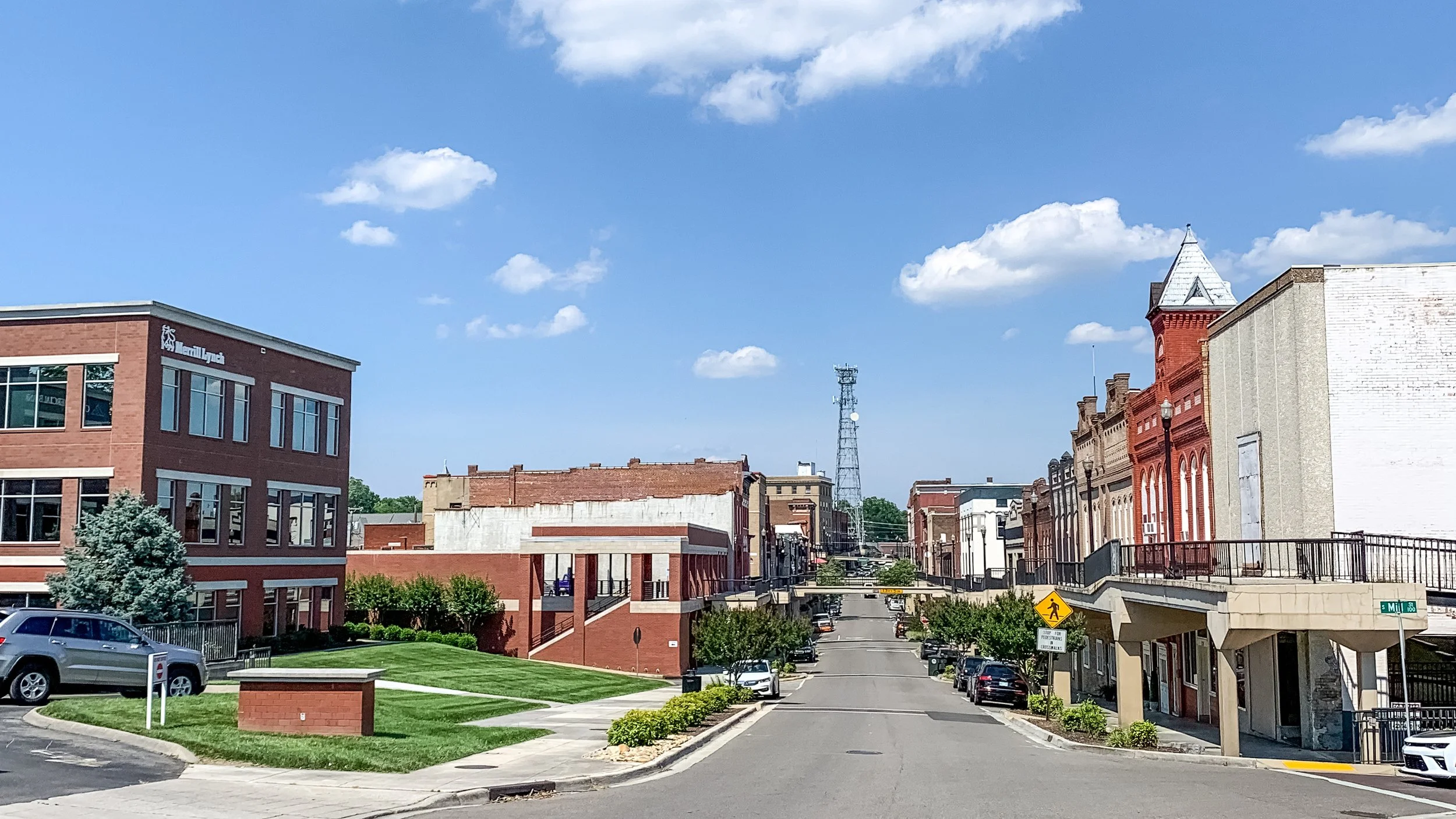 Downtown street scene with brick buildings, parked cars, a blue sky with clouds, and a noticeable radio tower in the background.