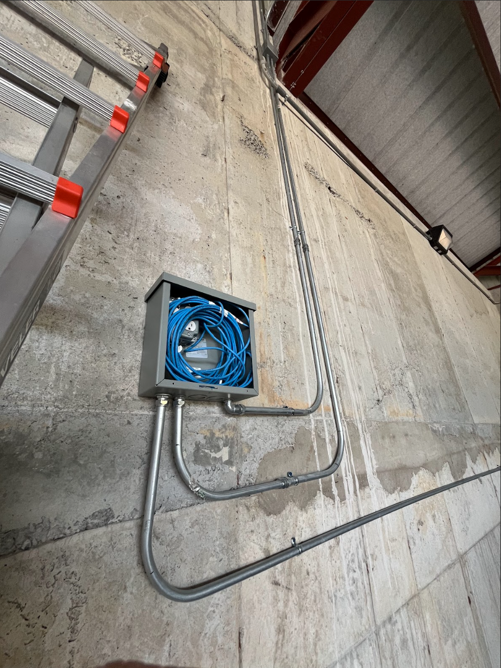 Electrical conduit and cable box mounted on a concrete wall with a metal ladder nearby.