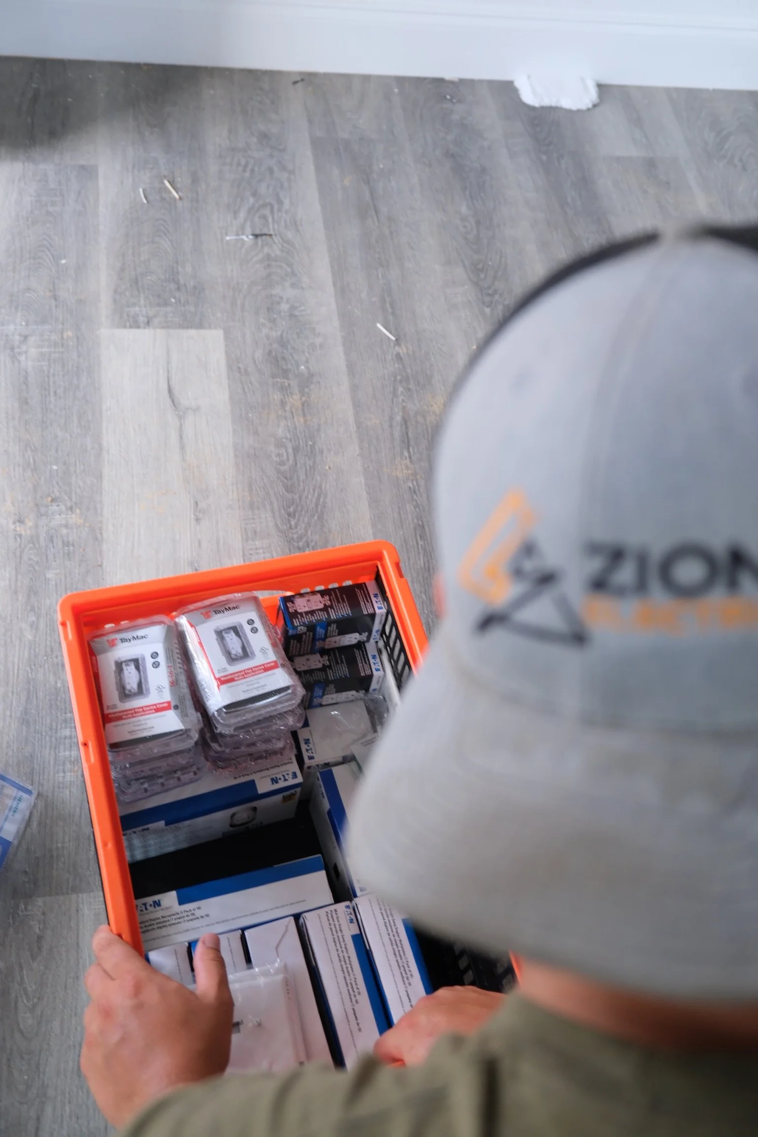 A person wearing a gray cap with an orange and black logo, looking into an orange container filled with various boxed and packaged items, on a gray wooden floor.