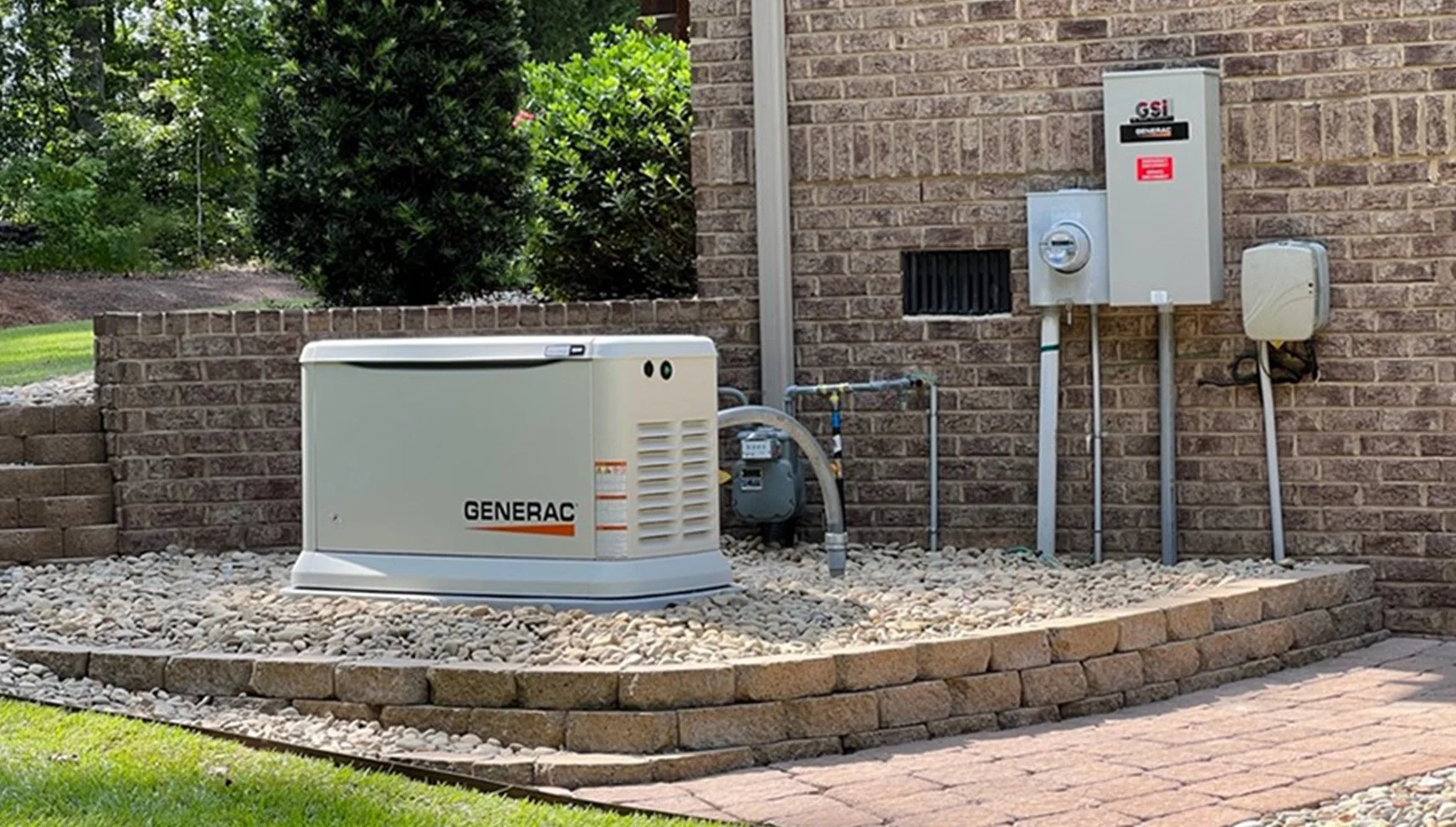 Outdoor view of a brick house's backyard corner with a Generac backup generator, electrical meter, and panel, surrounded by gravel and bordered by bricks, with green grass and trees in the background.
