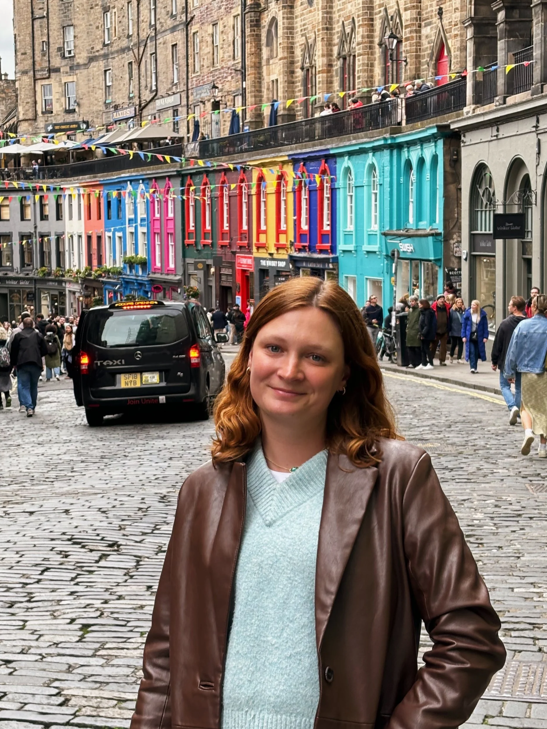 A woman with curly red hair wearing a brown leather jacket and a light blue sweater standing on a cobblestone street with colorful buildings and people in the background.