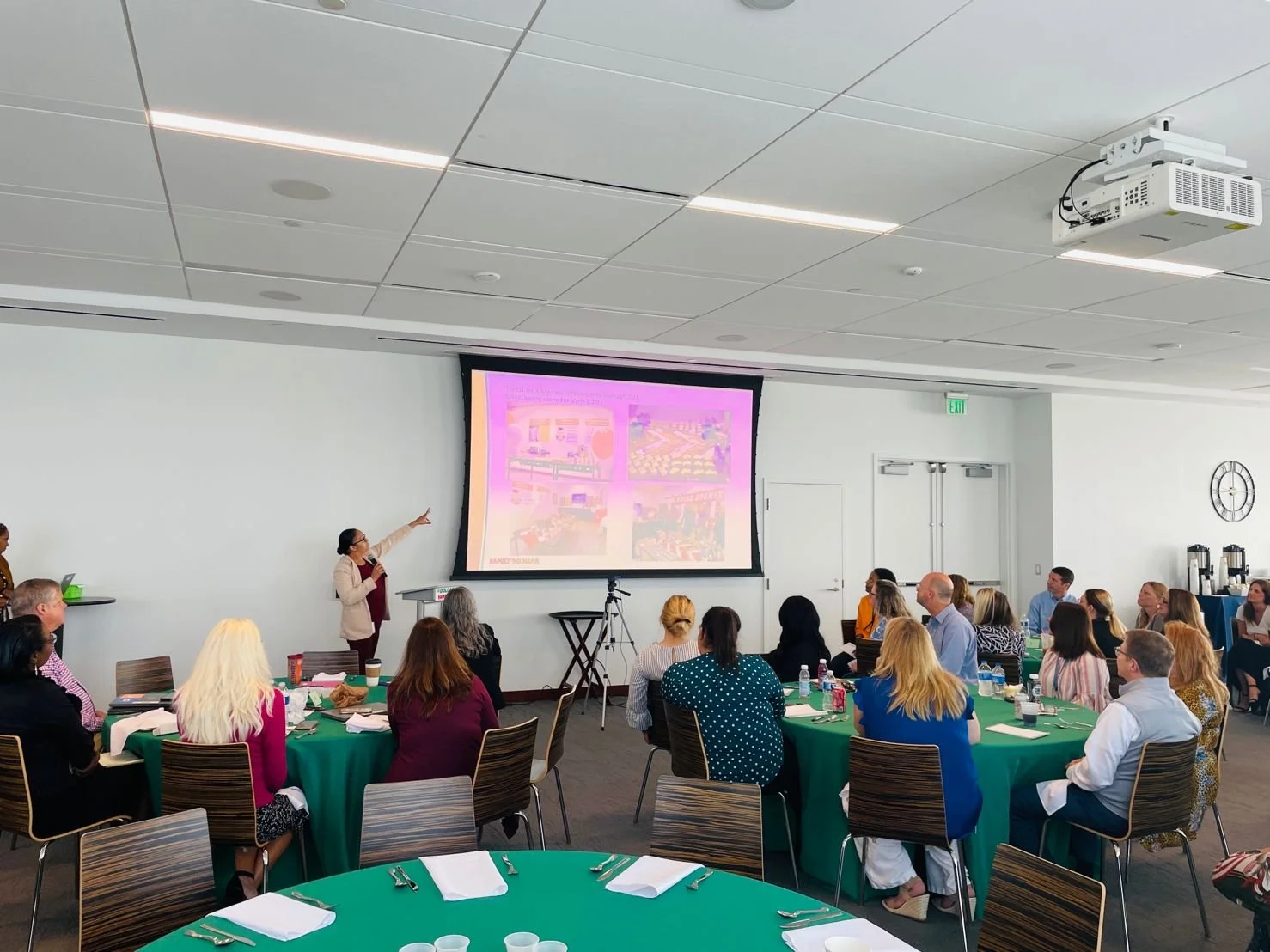 A woman is giving a presentation in a conference room with attendees seated at round tables. The screen behind her displays a slide with colorful images and text.