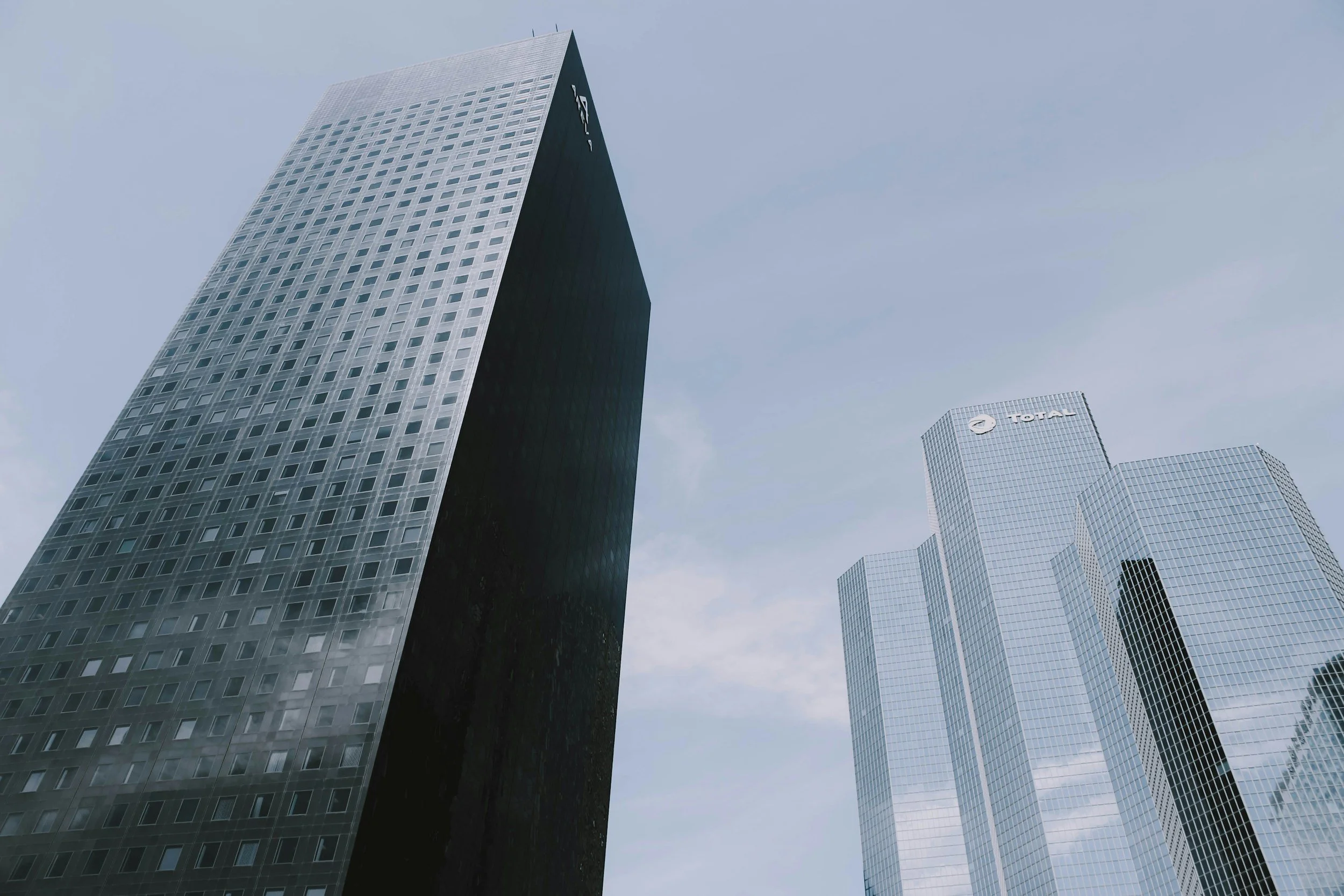 Modern skyscrapers with glass facades in a city skyline, one with a black exterior and another with a light-colored exterior, against a cloudy sky.