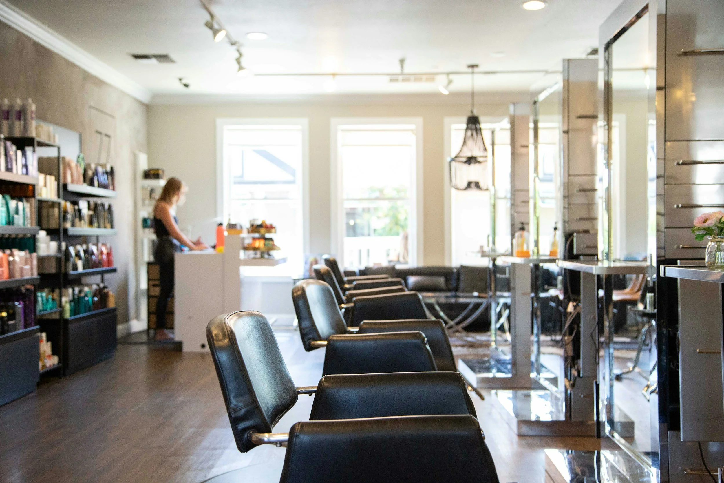 Empty hair salon with black chairs arranged in a row, shelves with hair products on the left, and a stylist working at the back, illuminated by large windows and hanging lights.