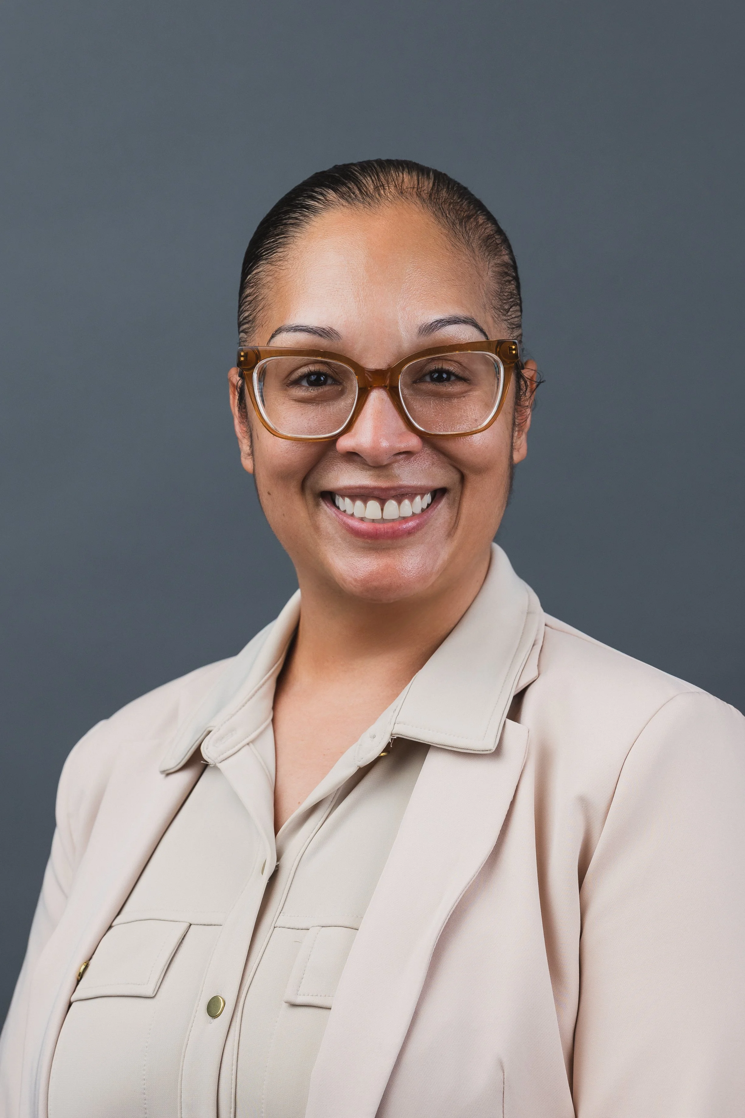 Professional woman smiling, wearing glasses and a beige blazer, against a plain gray background.