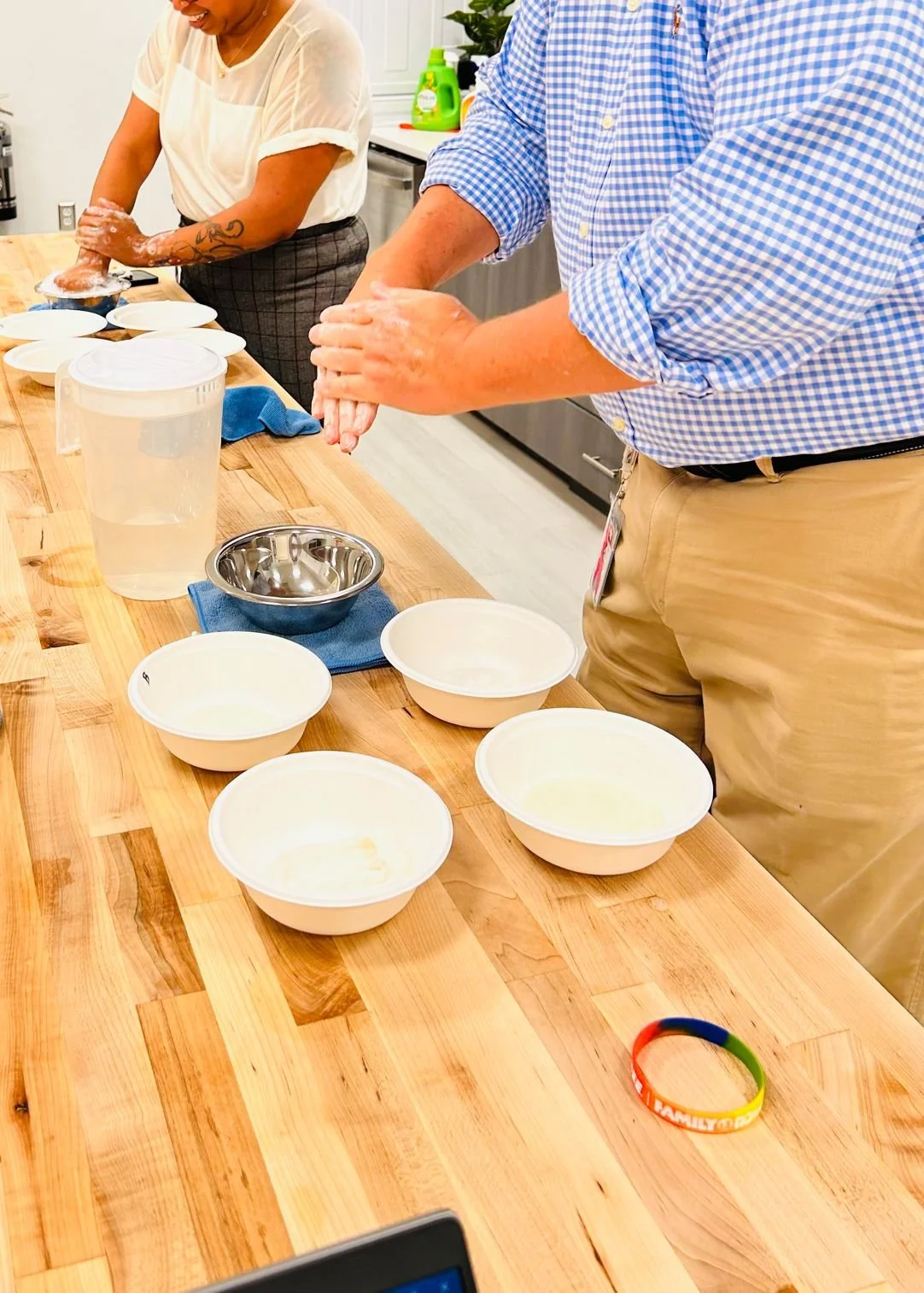 Two people are working together to make soap, kneading white soap mixture in their hands. There are multiple bowls with soap, a water pitcher, and a metal bowl on a wooden countertop, with a colorful rubber bracelet nearby.