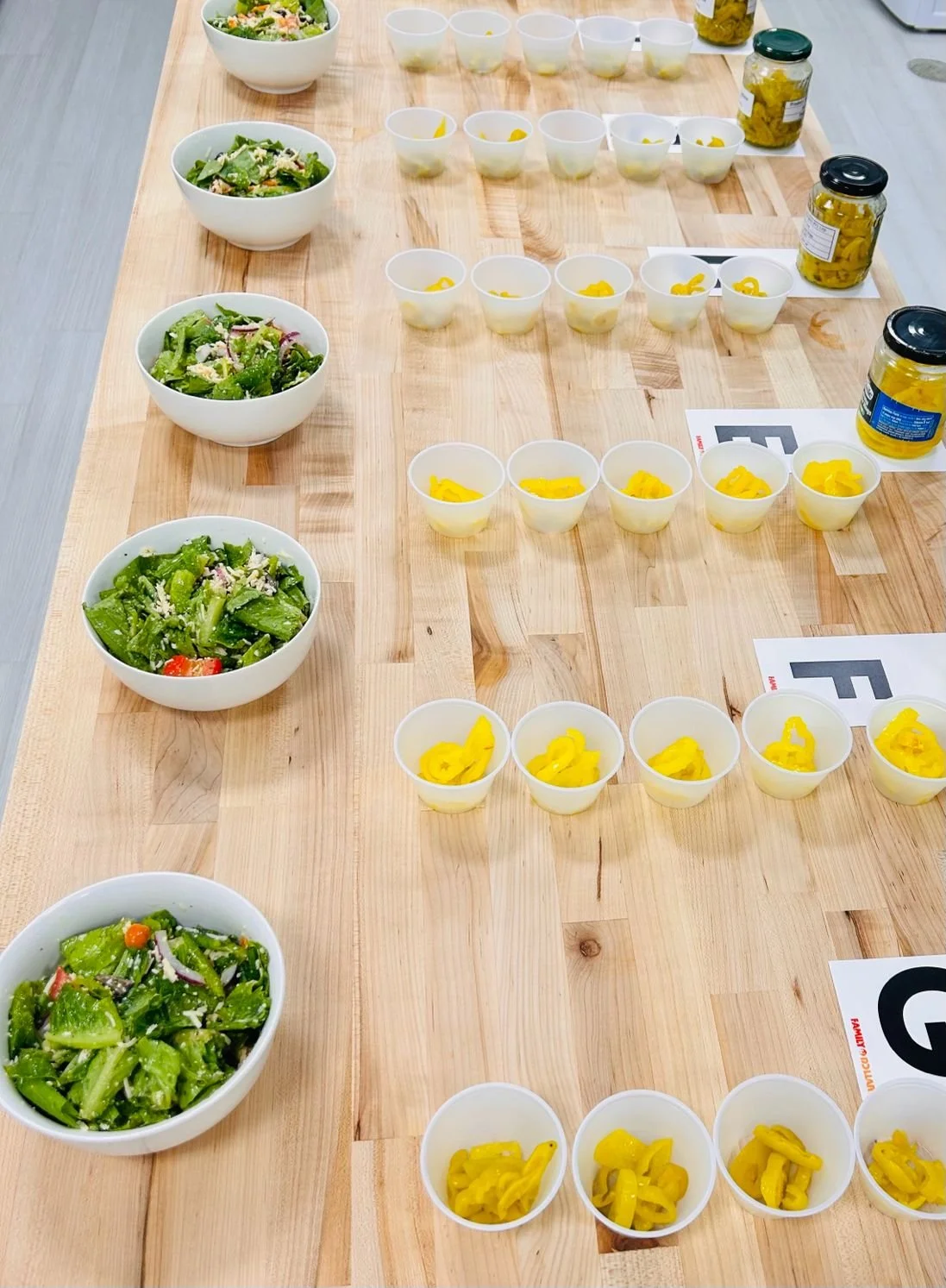 Arrangement of four bowls of mixed salad, each with different ingredients, and multiple small cups containing yellow pickled vegetables, on a light wooden table.