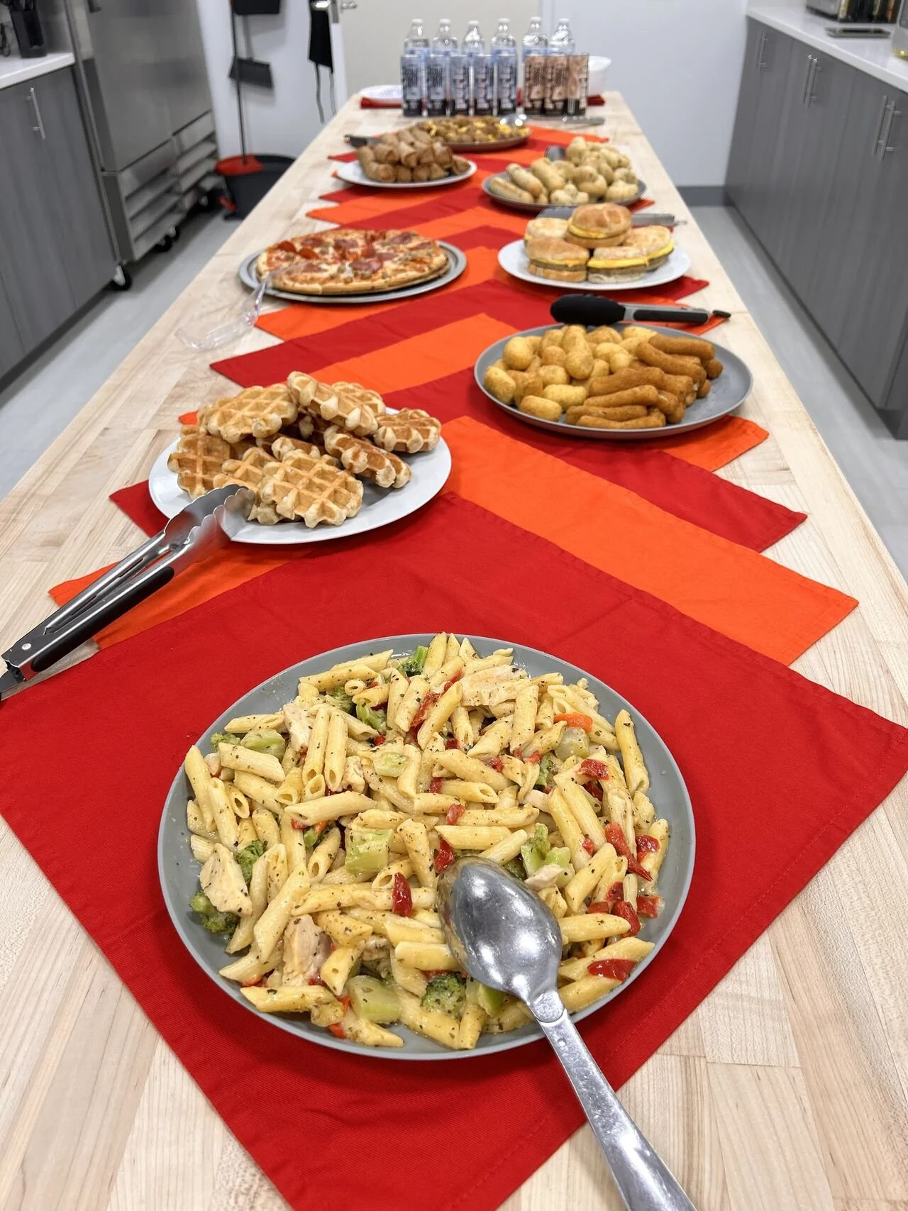 A long wooden table with a red and orange table runner, set with various foods including pasta salad, waffles, chicken nuggets, bread rolls, pizza, and rolls. There are also bottles of water at the far end.