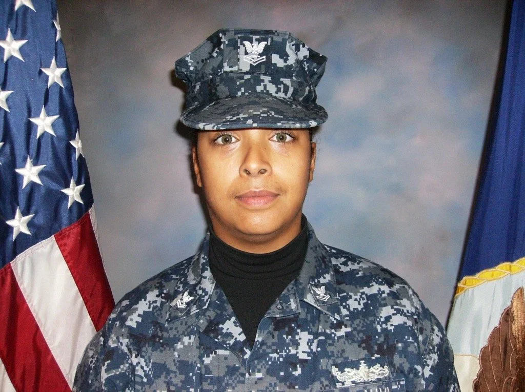 Female military officer in uniform standing in front of American and flag, wearing a camouflage hat and uniform with rank insignia, in front of a backdrop with cloudy sky pattern.