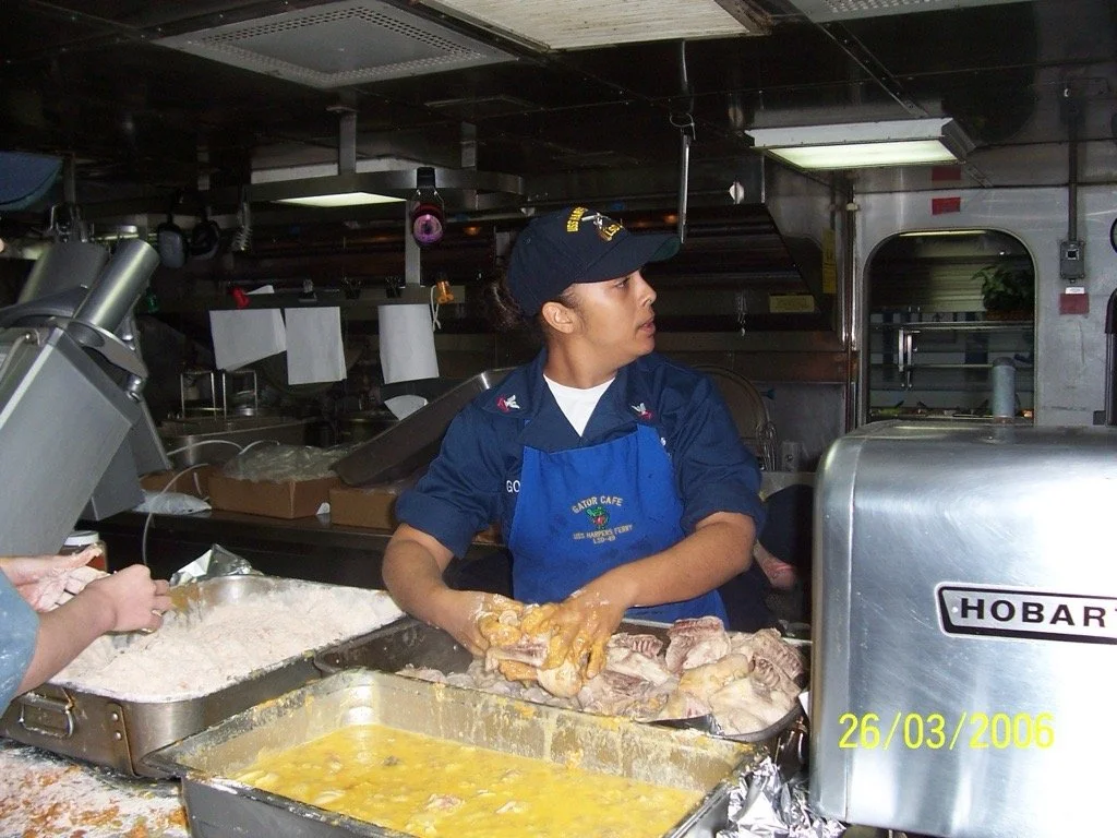 A woman in a uniform and apron working in a kitchen, handling raw meat and eggs, with cooking equipment and kitchen items around.