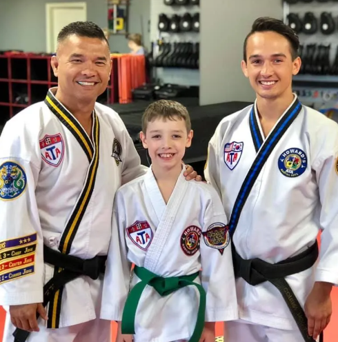 Three males in martial arts uniforms standing in a gym, smiling at the camera. The boy in the middle has a green belt, and the two men have black belts. The background shows training equipment and a red padded floor.