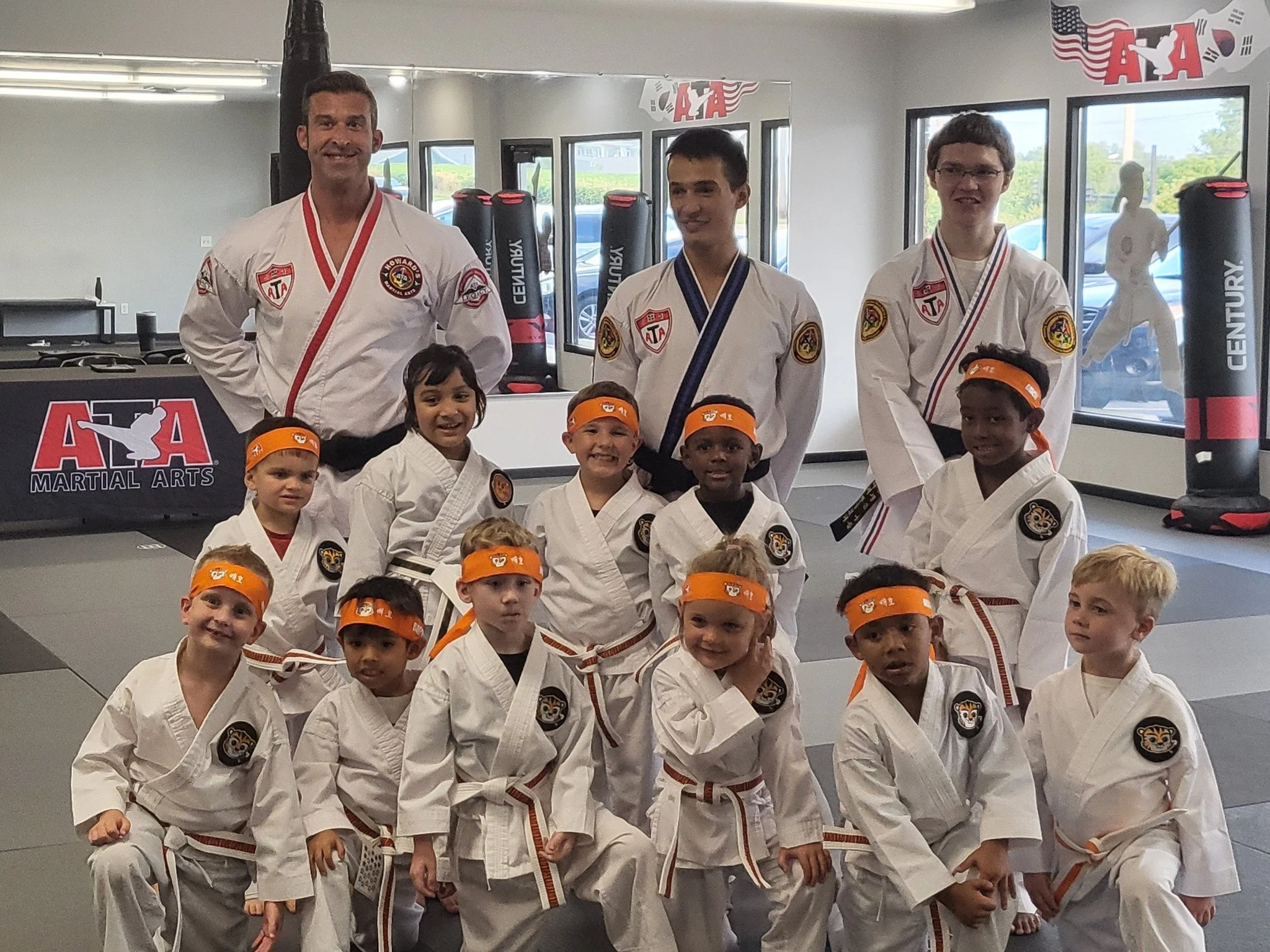 Group of children and adults in martial arts uniforms in a martial arts studio, with some children wearing orange headbands, posing for a photo.