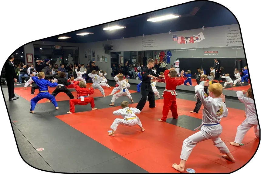 Children practicing martial arts in a dojo or martial arts training center with an instructor, wearing colored uniforms, in front of a mirror and a wall with martial arts gear and flags.
