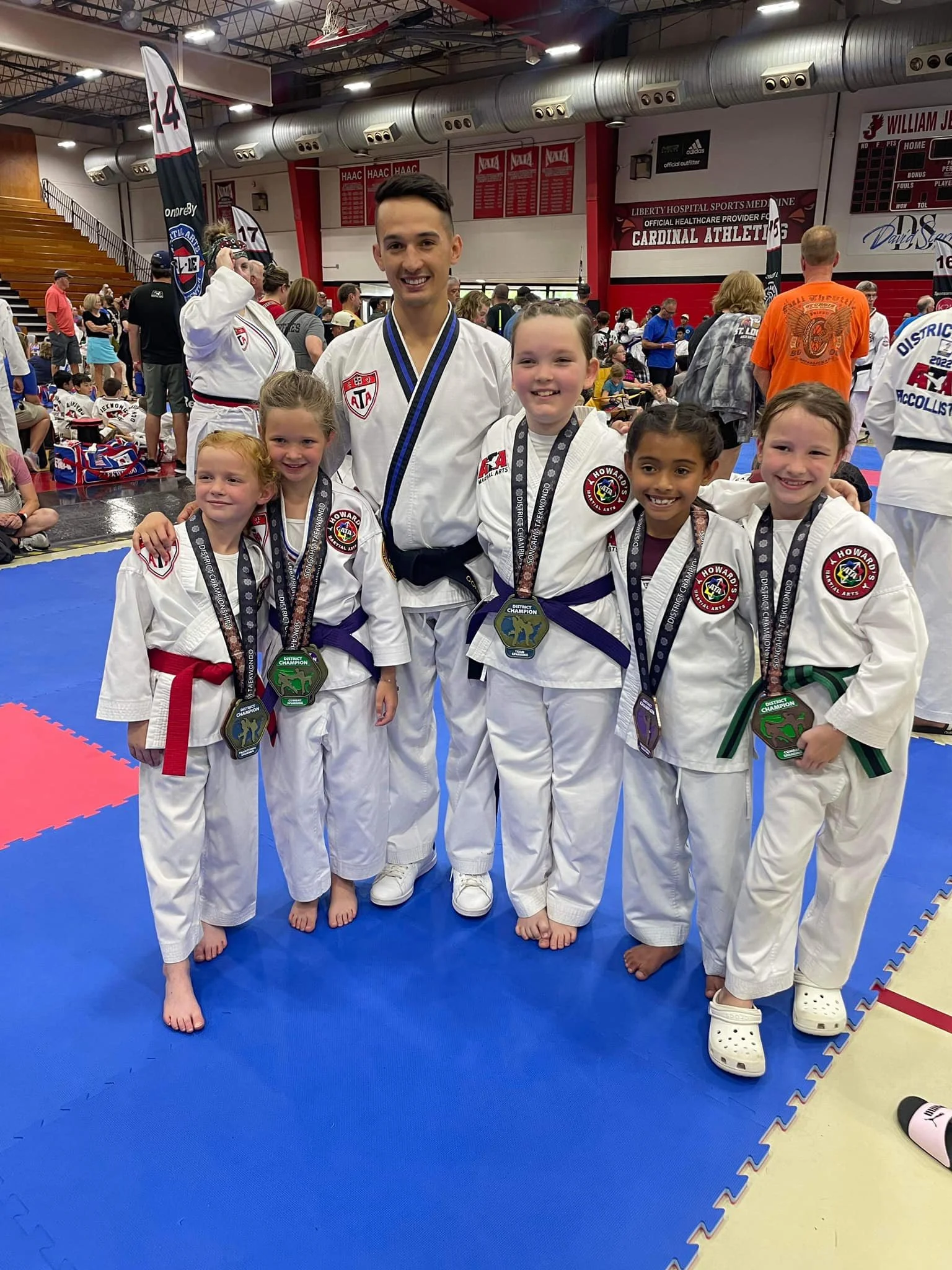 Group of young girls and their instructor posing with medals after a martial arts tournament, inside a gymnasium with banners hanging from the ceiling.