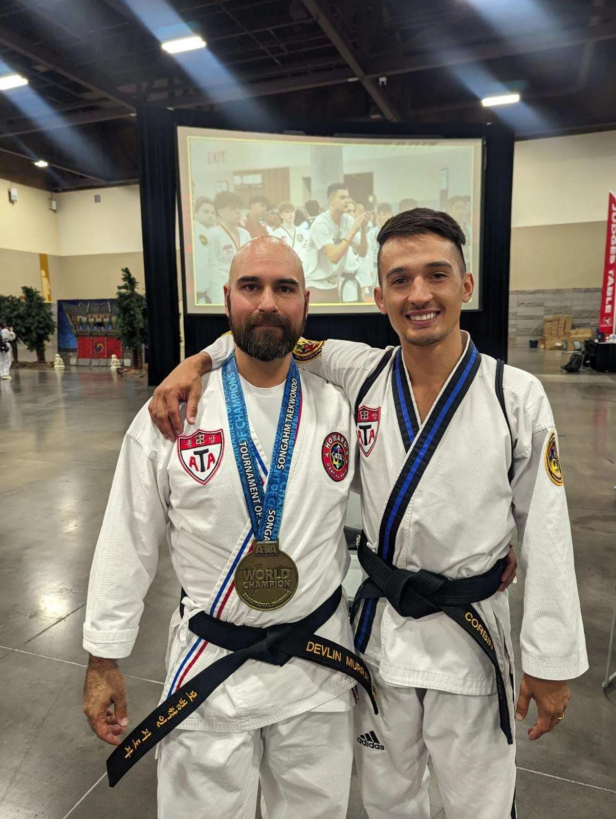 Two men in martial arts uniforms smiling, one with a gold medal around his neck, standing indoors with a large screen in the background showing a group of people, some in martial arts uniforms.