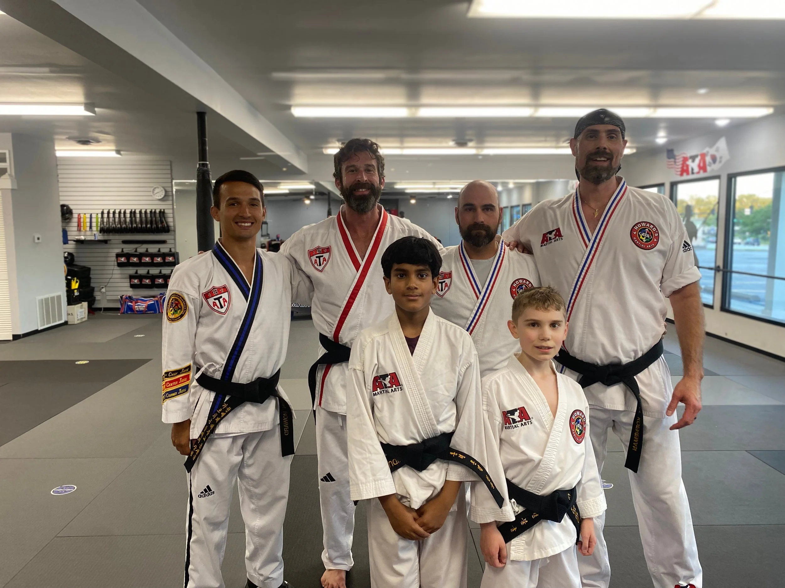 Group of five men and two boys in martial arts uniforms standing inside martial arts training center.