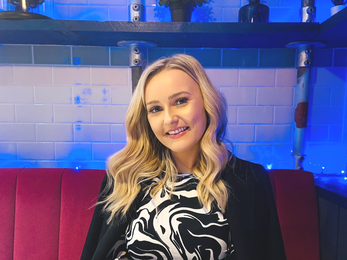 A young woman with blonde wavy hair sitting on a red cushioned bench in a room with blue lighting, white brick walls, and a black shelf above her.